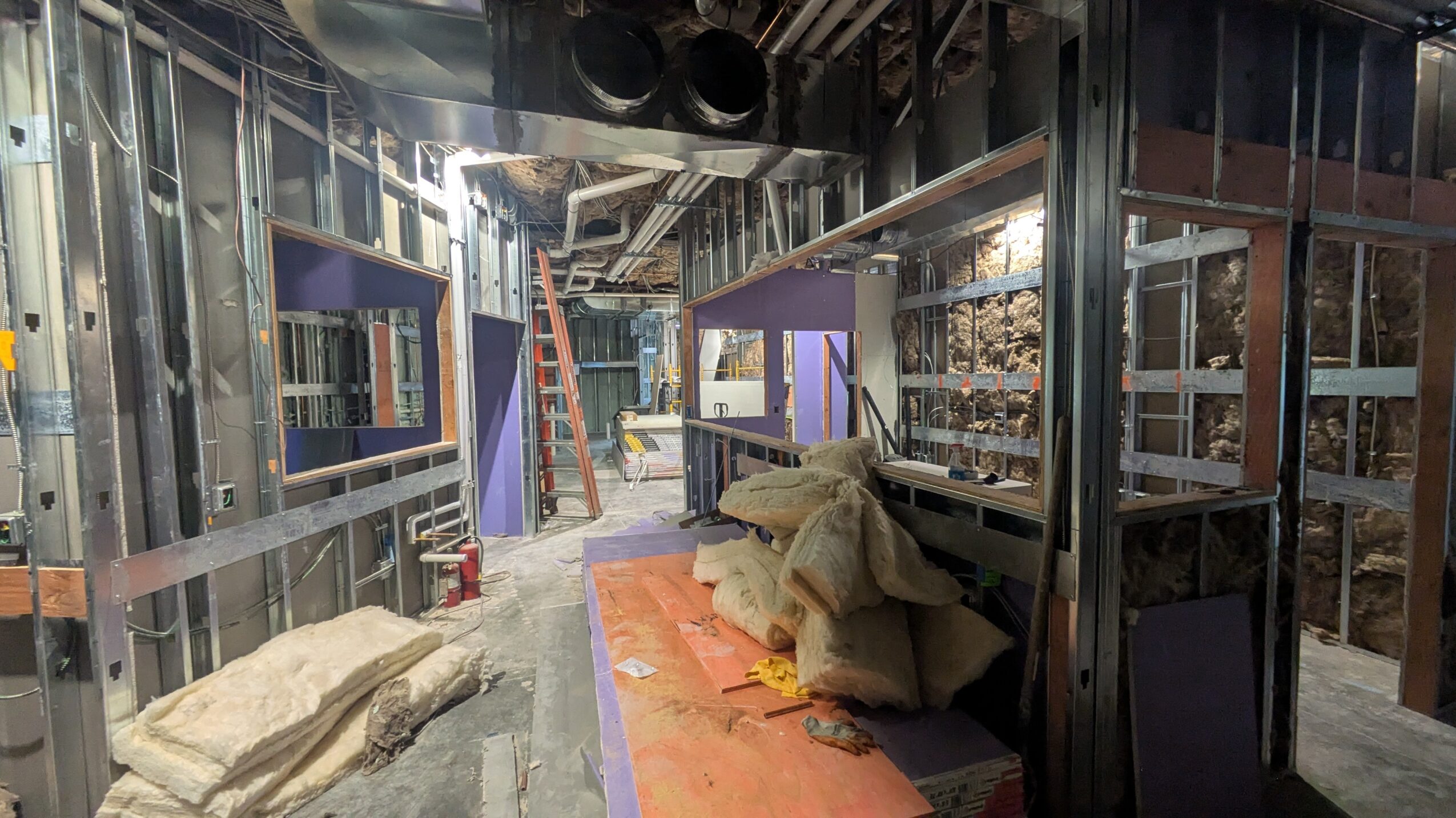 Construction site with metal framing, tools, and materials, including bags and a ladder, in an unfinished building interior.