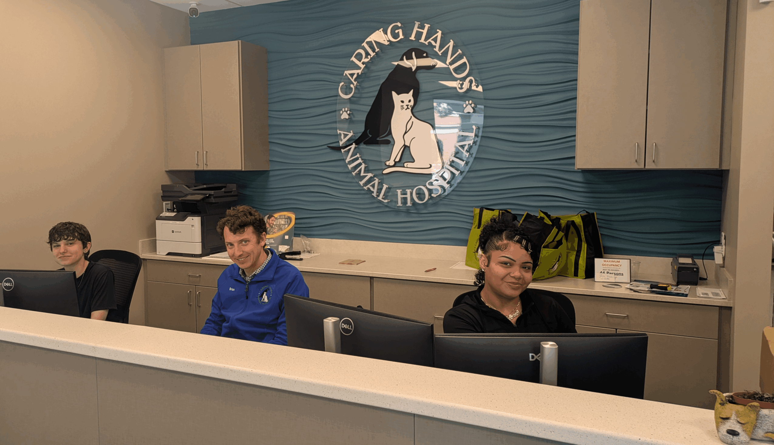 Three people sit at a desk in an animal shelter reception area, with a sign reading 'Caring Hands Animal Hospital' behind them.