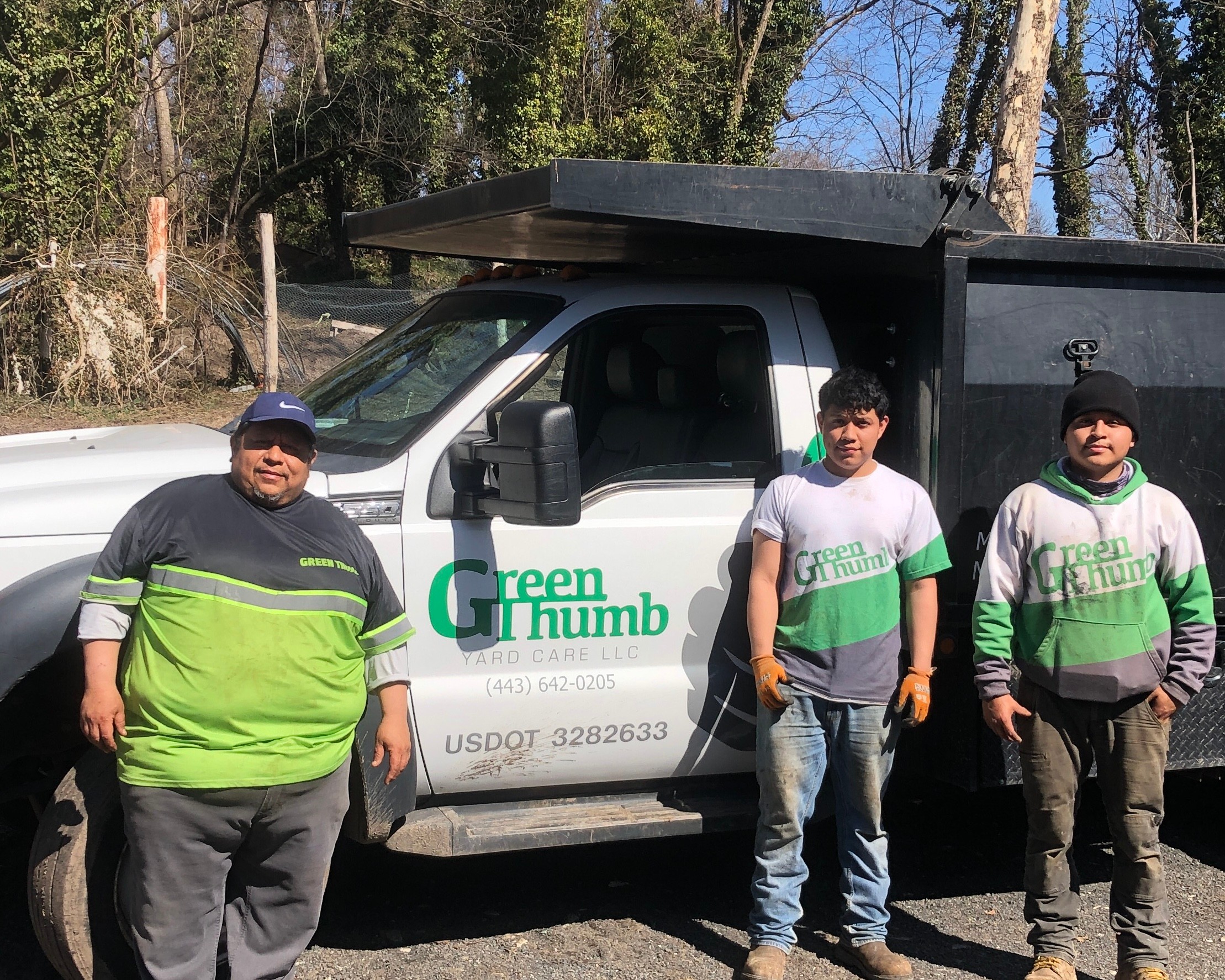 Three people standing outdoors in front of a truck with a green and white logo, trees in the background.