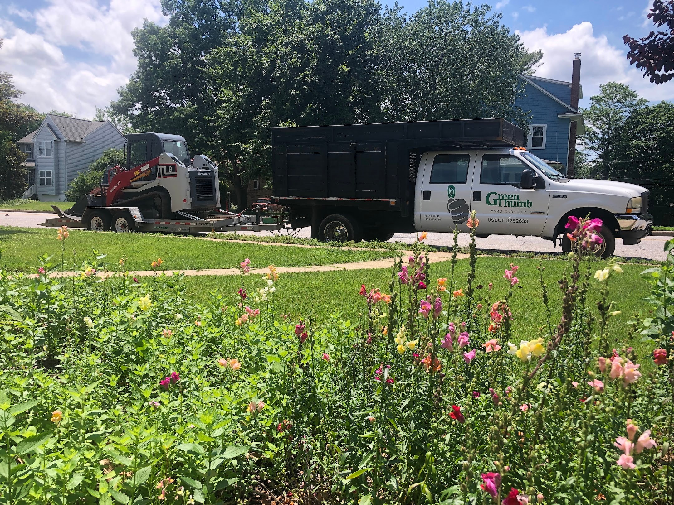 Pickup truck with a black bed and a white cab parked on a residential street, with a tow truck nearby, and a garden with pink flowers in foreground.