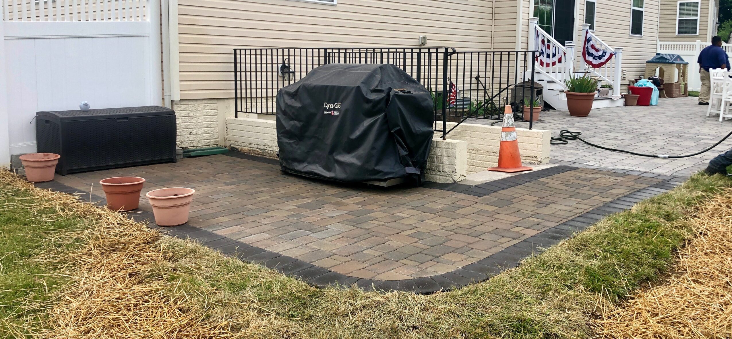 Backyard patio with a covered grill, potted plants, and a black bench, bordered by grass and a house with a porch.