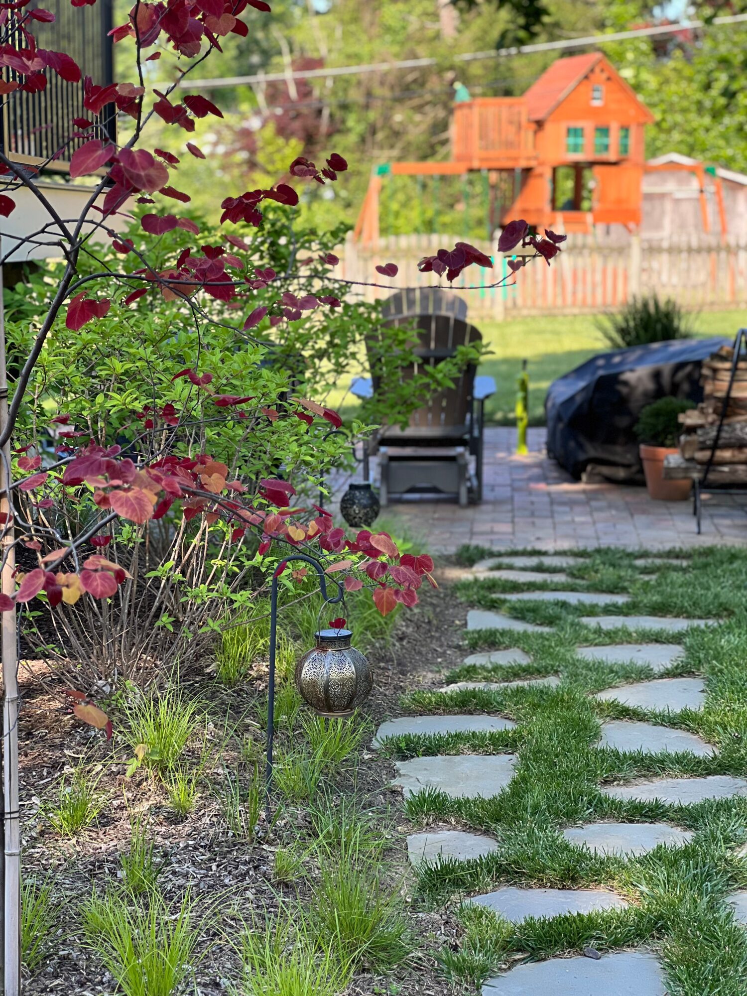 Path with stepping stones leads to a park bench, with a car and a house in the background, surrounded by greenery.