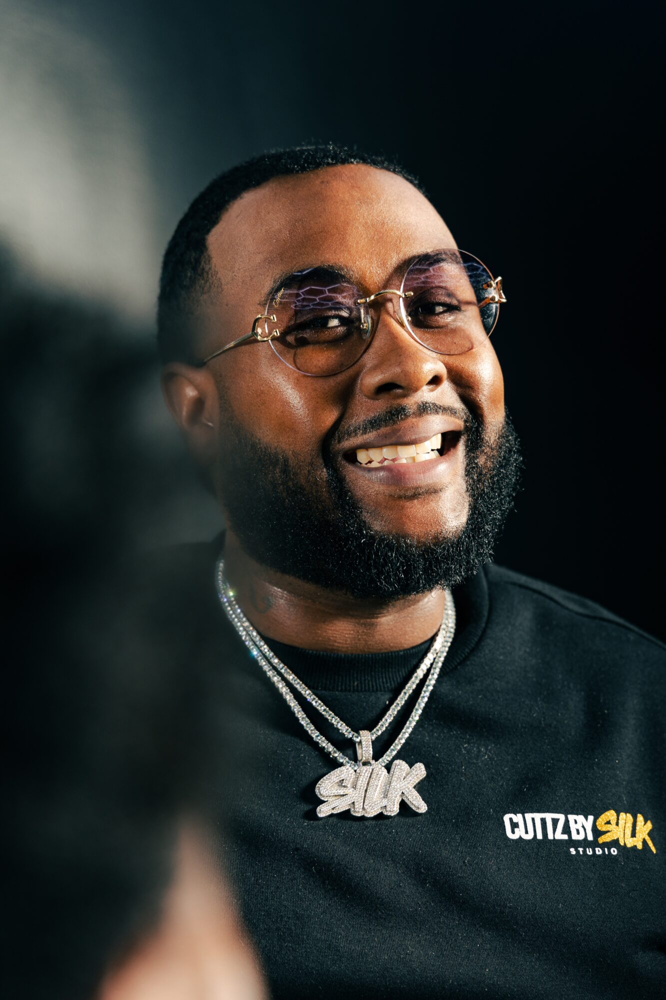 Smiling man with glasses, beard, and jewelry, wearing a black shirt with a logo, against a dark background.