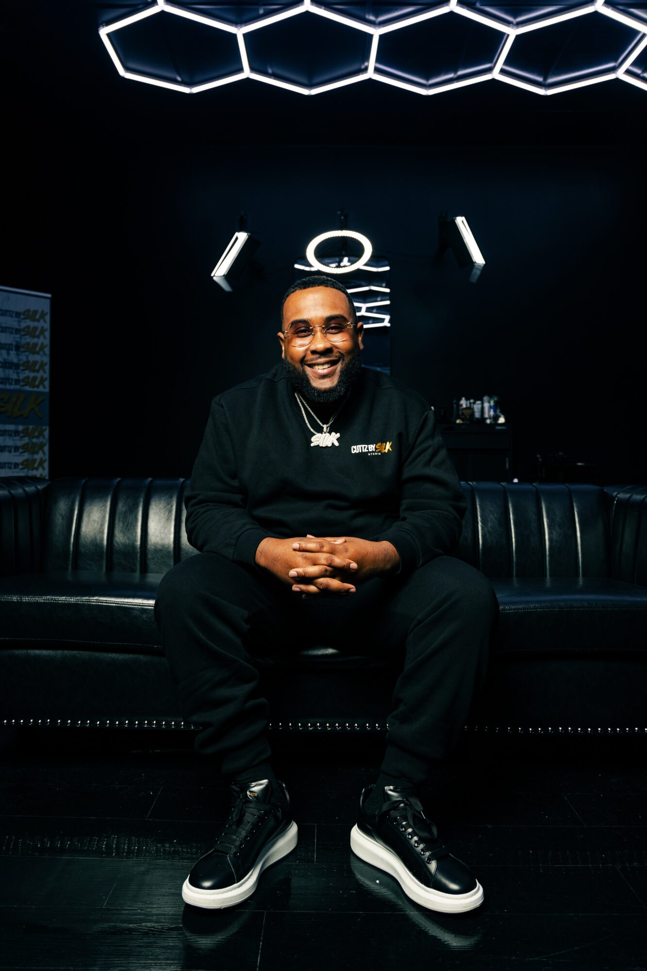 Person sitting on a black leather couch, smiling, in a dark room with illuminated symbols on the wall behind.