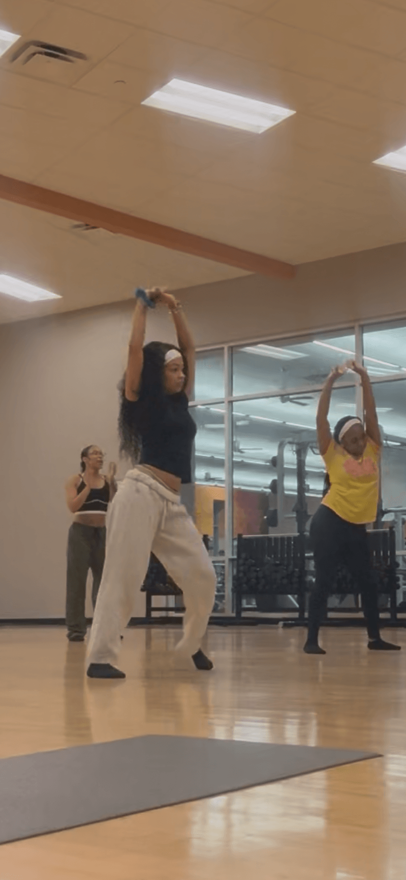 Three people in a dance studio with large windows, practicing dance moves with arms raised, wooden floor, ceiling lights, and a yoga mat.
