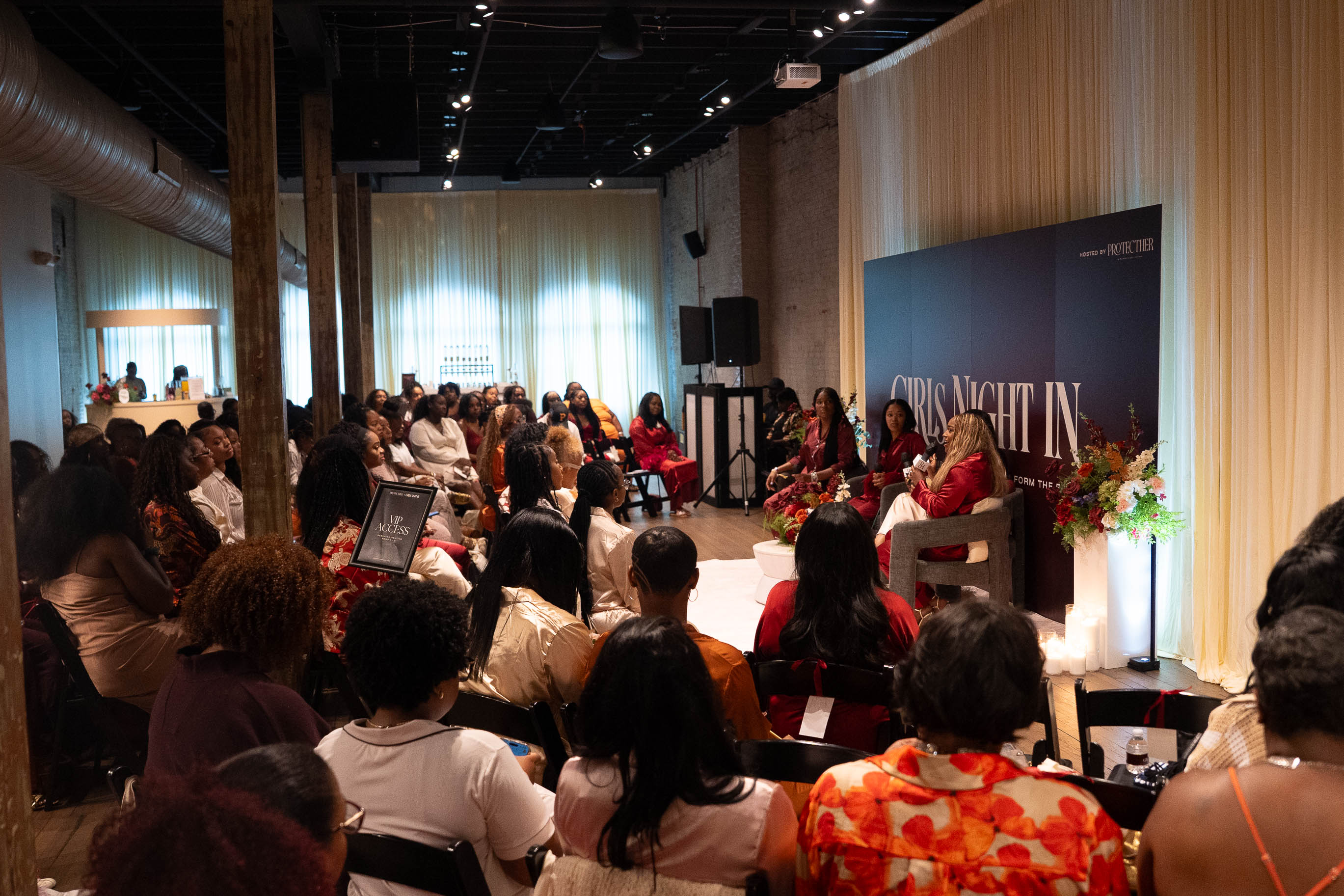 Panel discussion with five women on stage in front of an audience in a large room with wooden walls and curtains.
