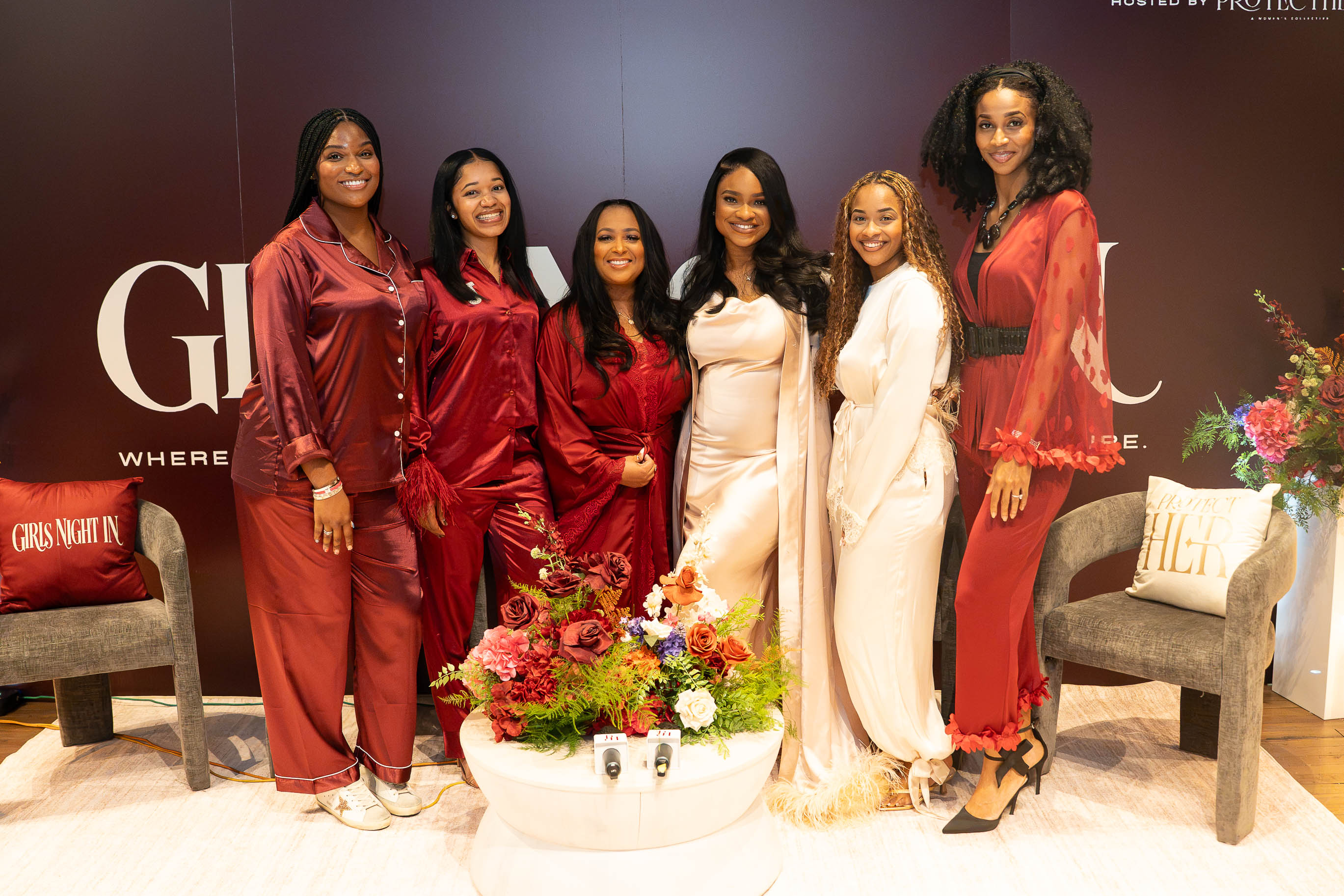 Six women in satin pajamas pose together indoors, with a flower arrangement in front and chairs on either side.