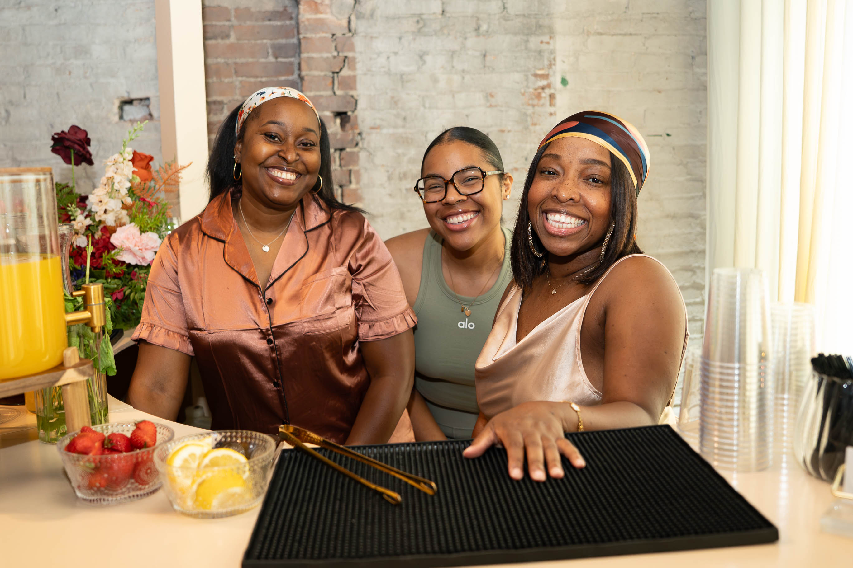 Three women smiling at a table with drinks and fruit, in a bright room with a brick wall and flowers.