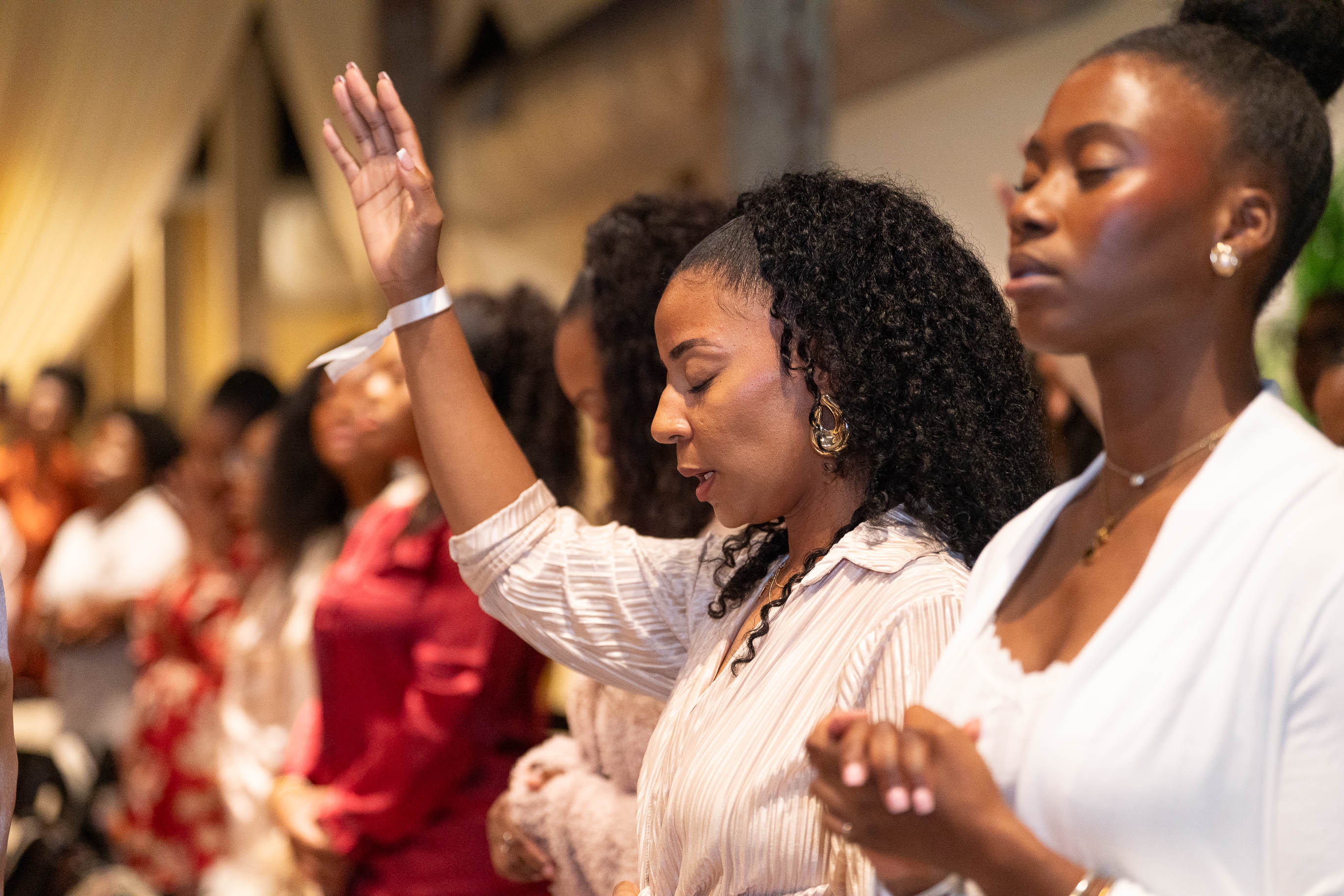 Two women with eyes closed, one raising her hand, in a group setting, possibly praying or meditating.