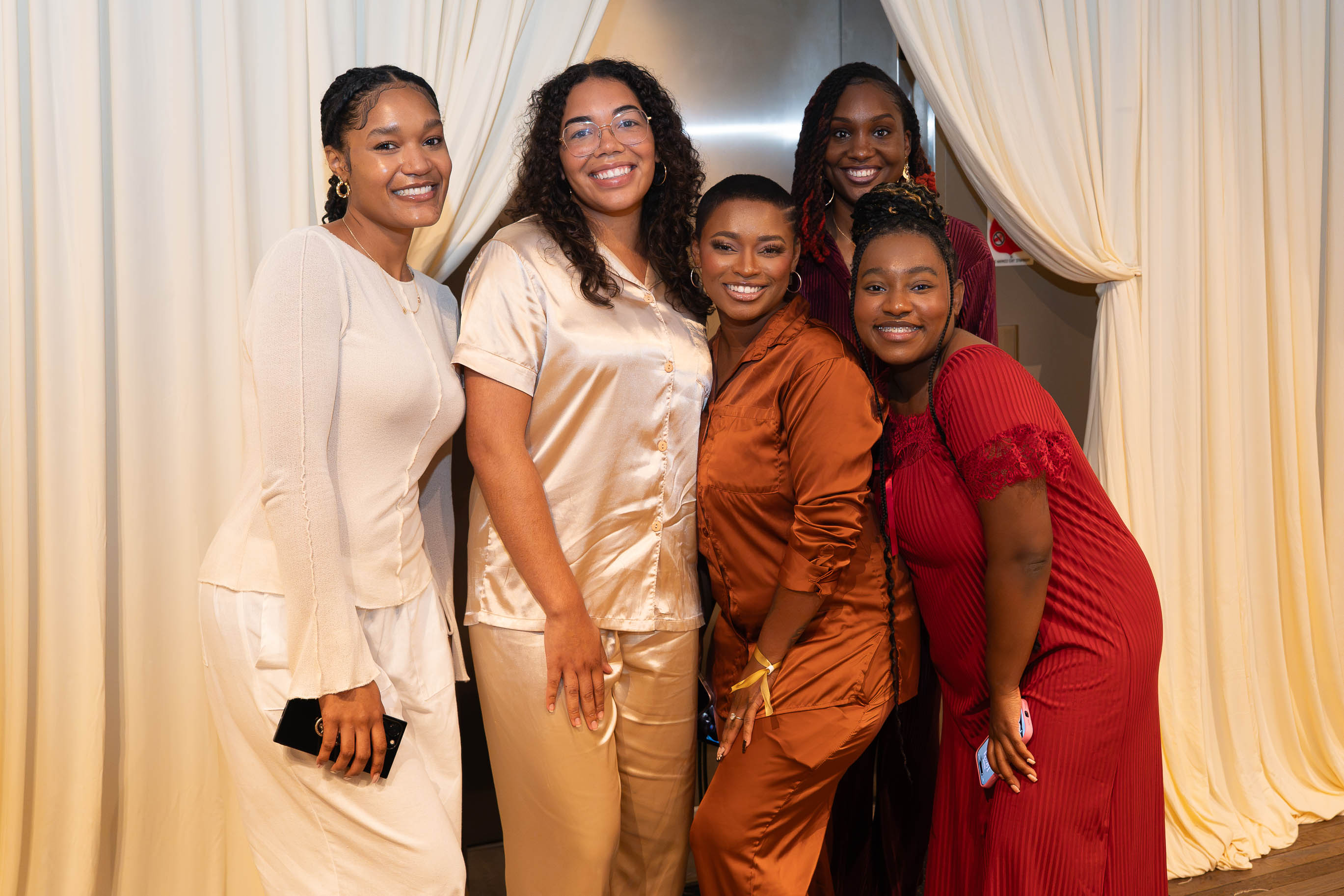Five women smiling and posing together indoors with cream-colored curtains in the background.