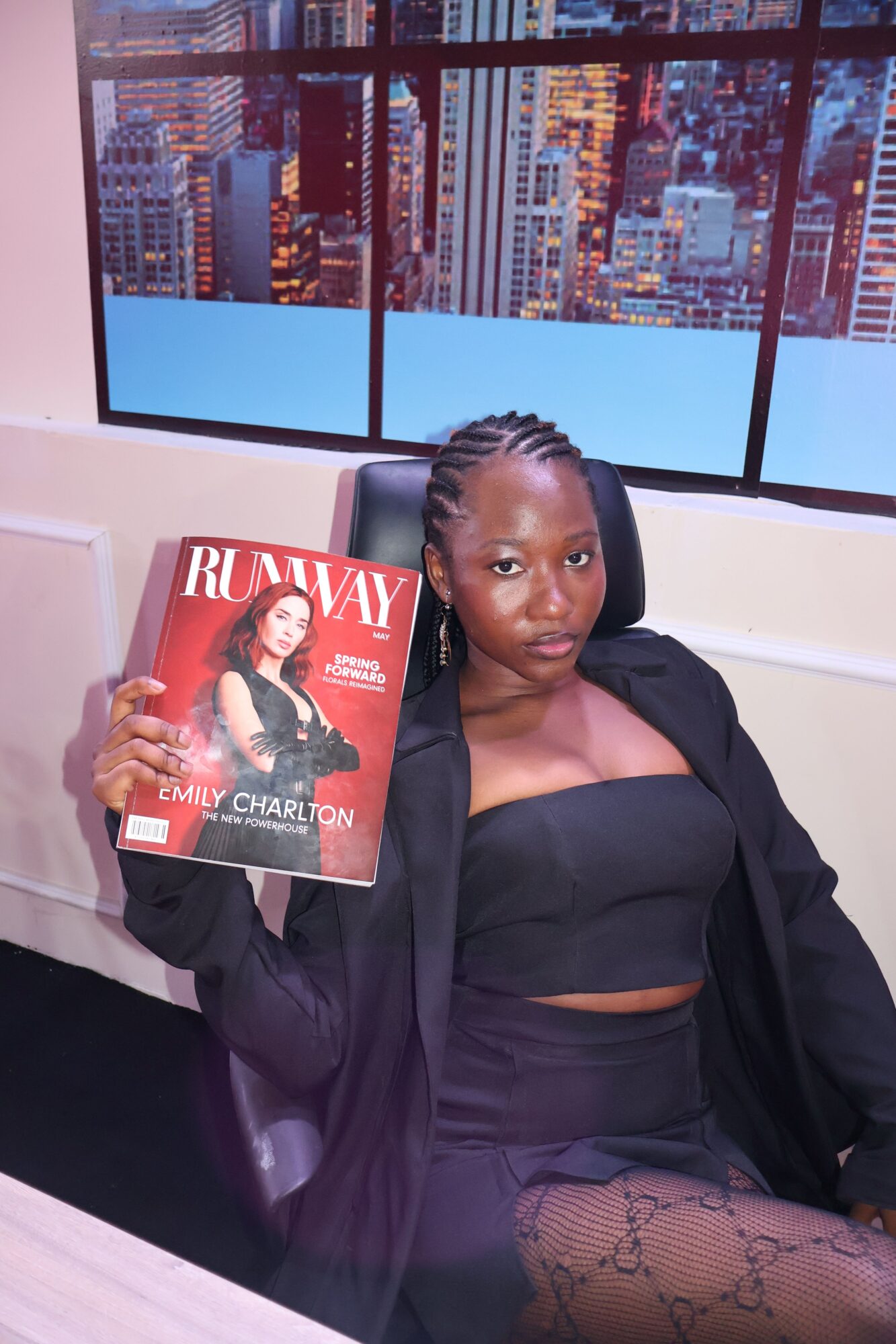 Woman in black outfit holding a magazine, sitting in front of a cityscape window, with a city skyline background.