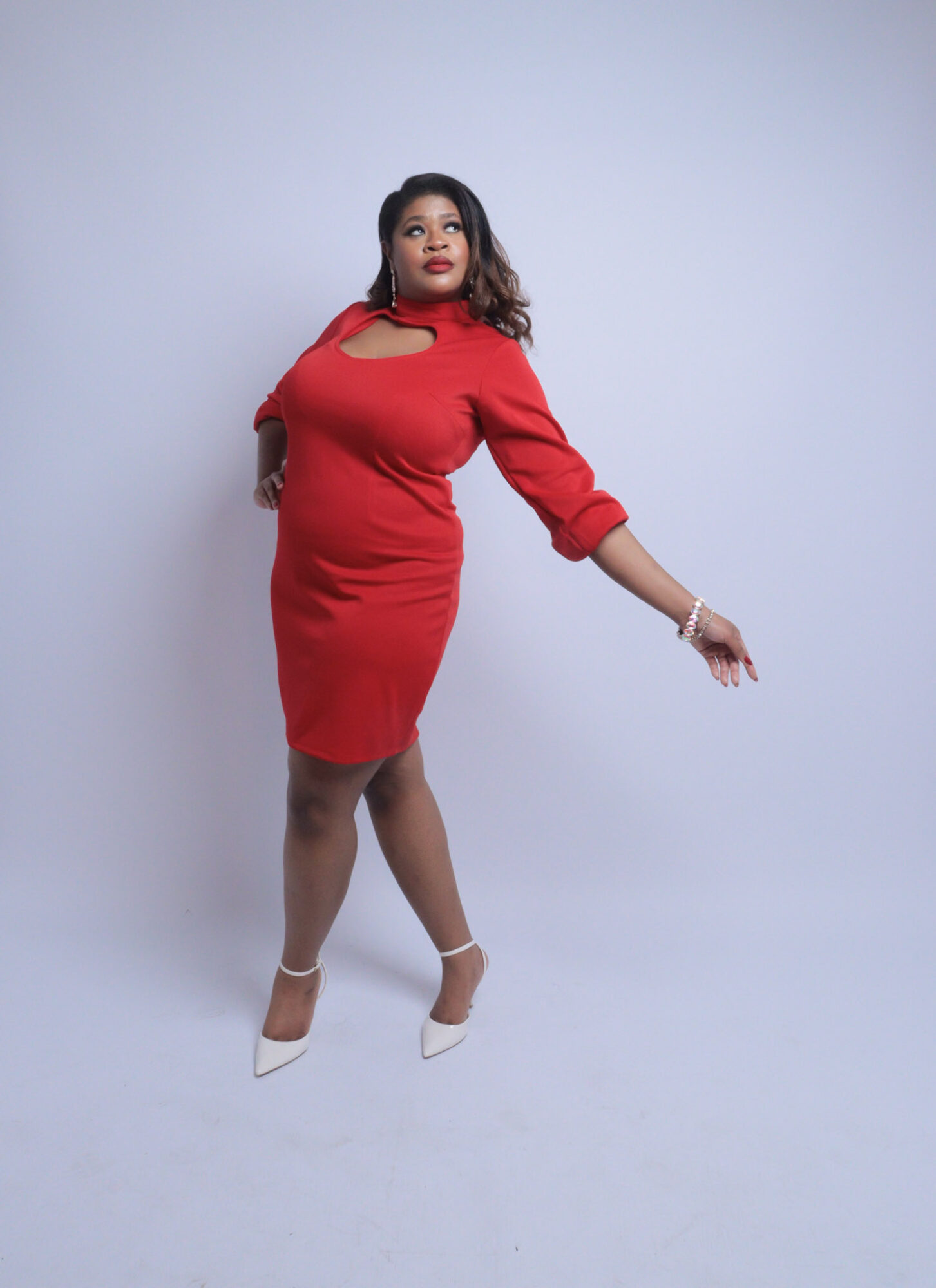 Woman in red dress standing against light background, posing with one arm extended, looking to the side.