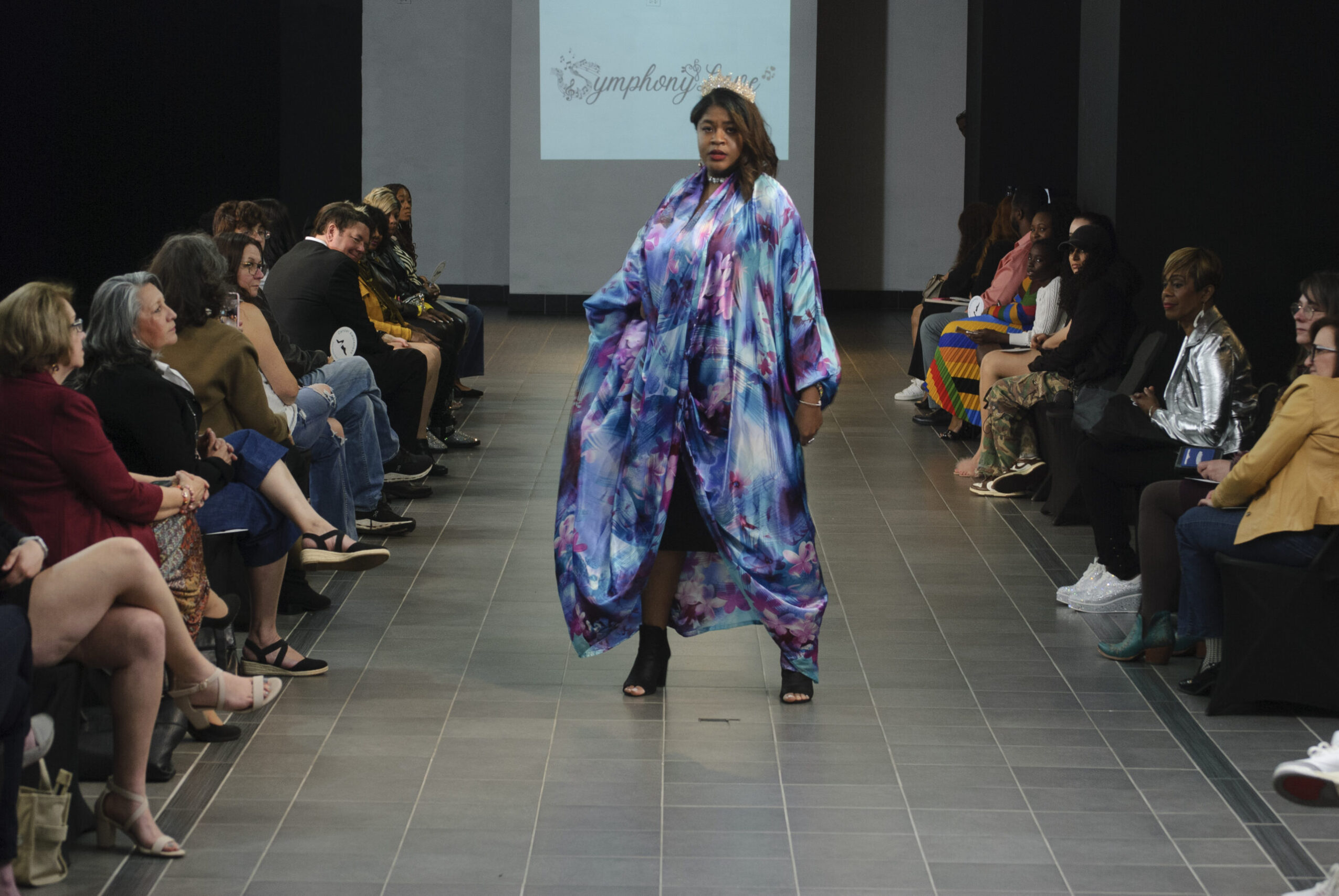 Model walking down runway in colorful, flowing dress with audience seated on both sides, background screen displaying text.