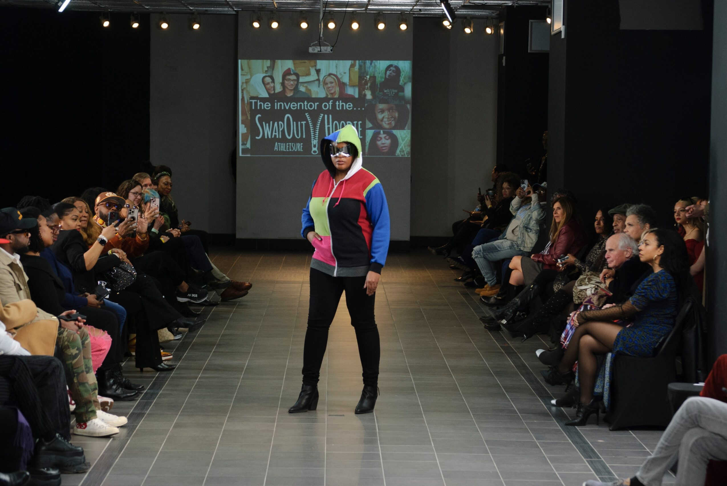Person walking on runway during fashion show with audience seated on both sides, and a screen in the background.