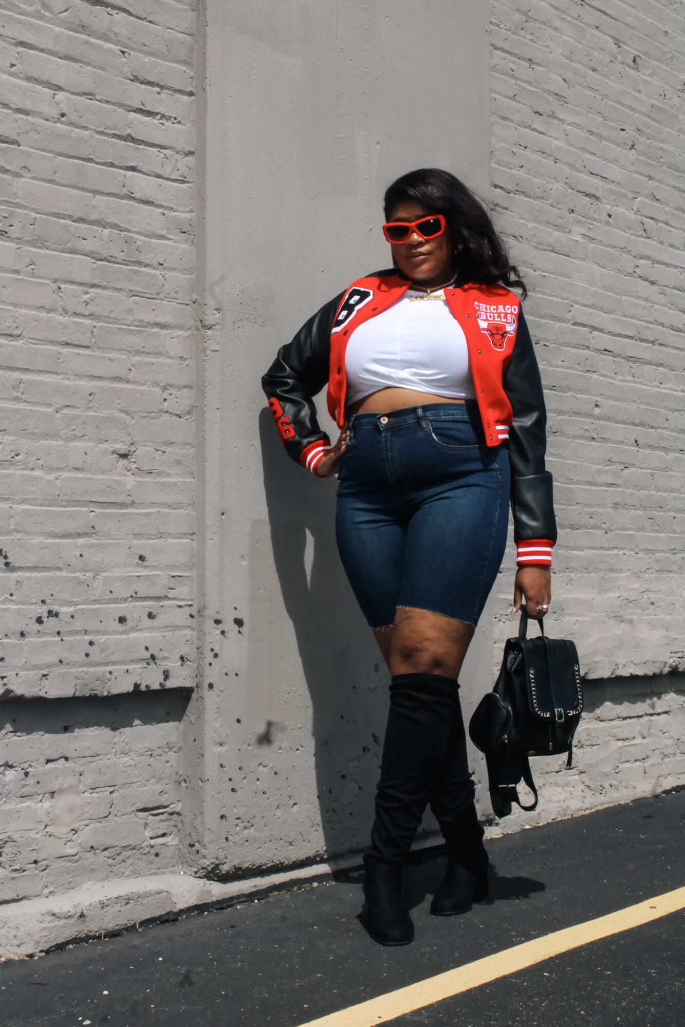 Woman with sunglasses and curly hair leaning against a wall, wearing a red and black jacket, white top, jeans, and thigh-high boots, holding a bag.