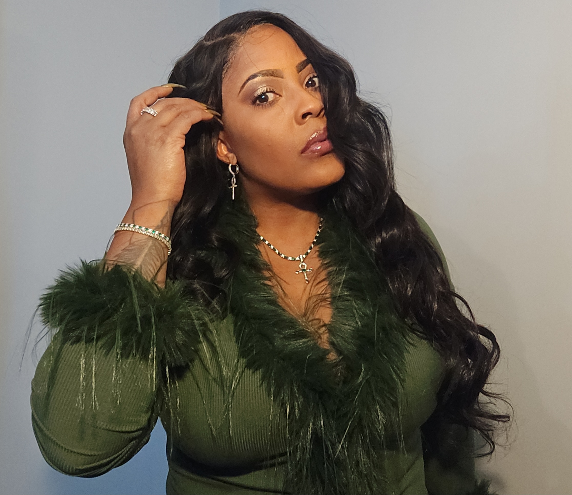 Woman with long wavy hair wearing a green dress with feathered sleeves, jewelry, and makeup, posing against a plain background.