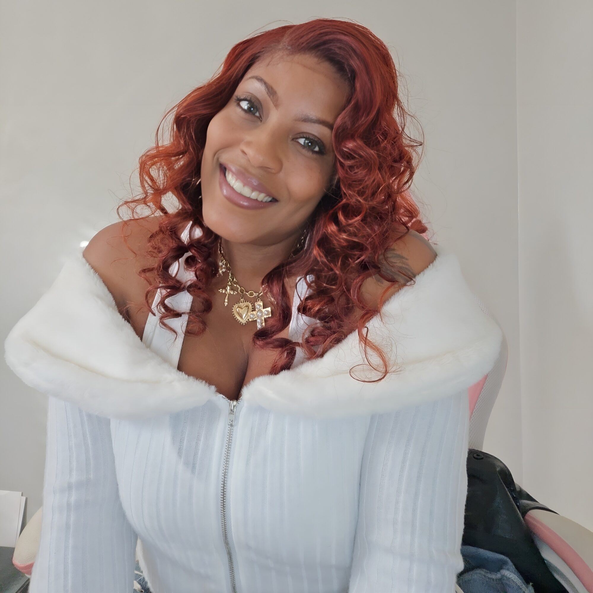 Smiling woman with red curly hair wearing a white zip-up top and a gold necklace, posing indoors.