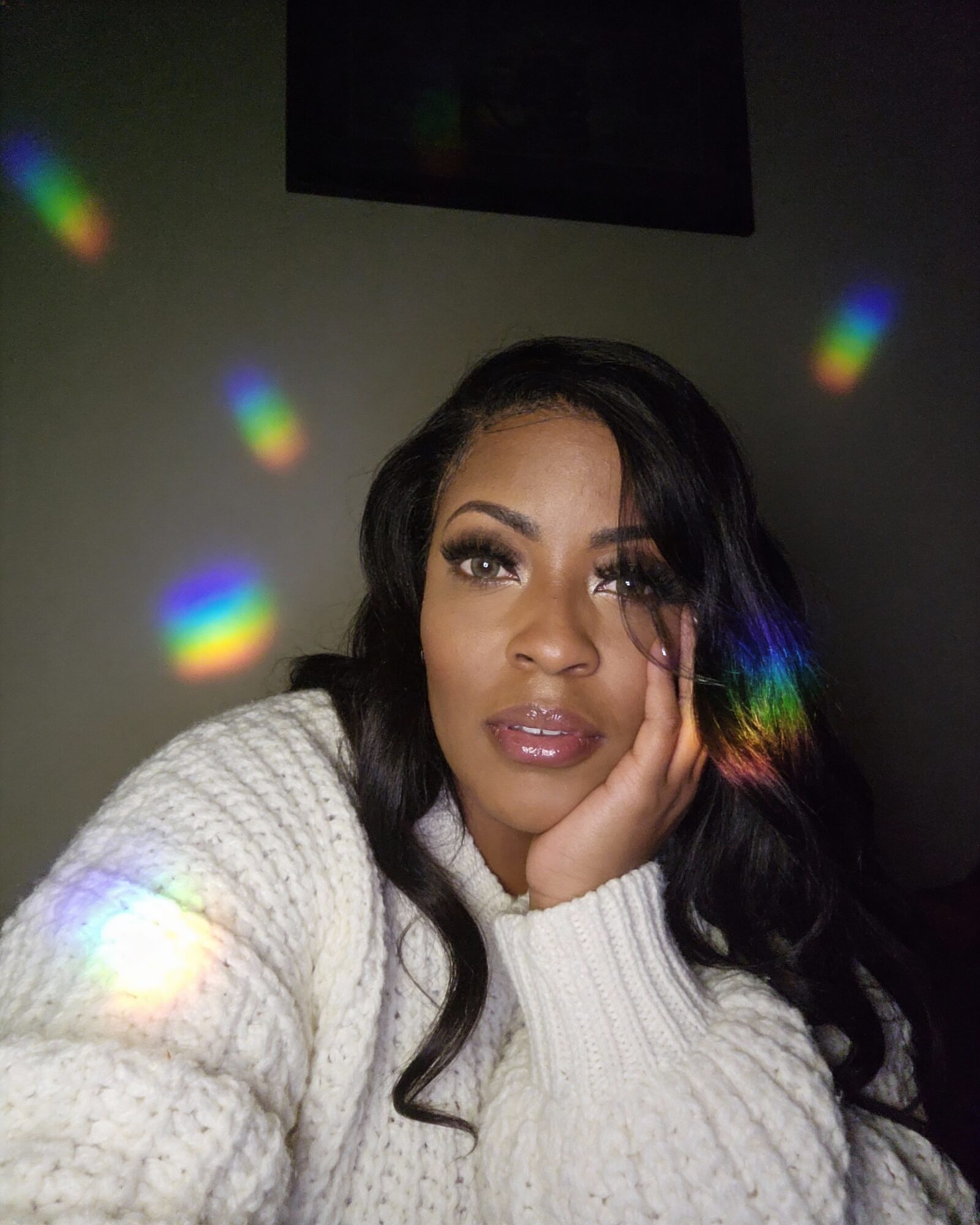 Woman with long dark hair and makeup, resting her face on her hand, surrounded by colorful light reflections.