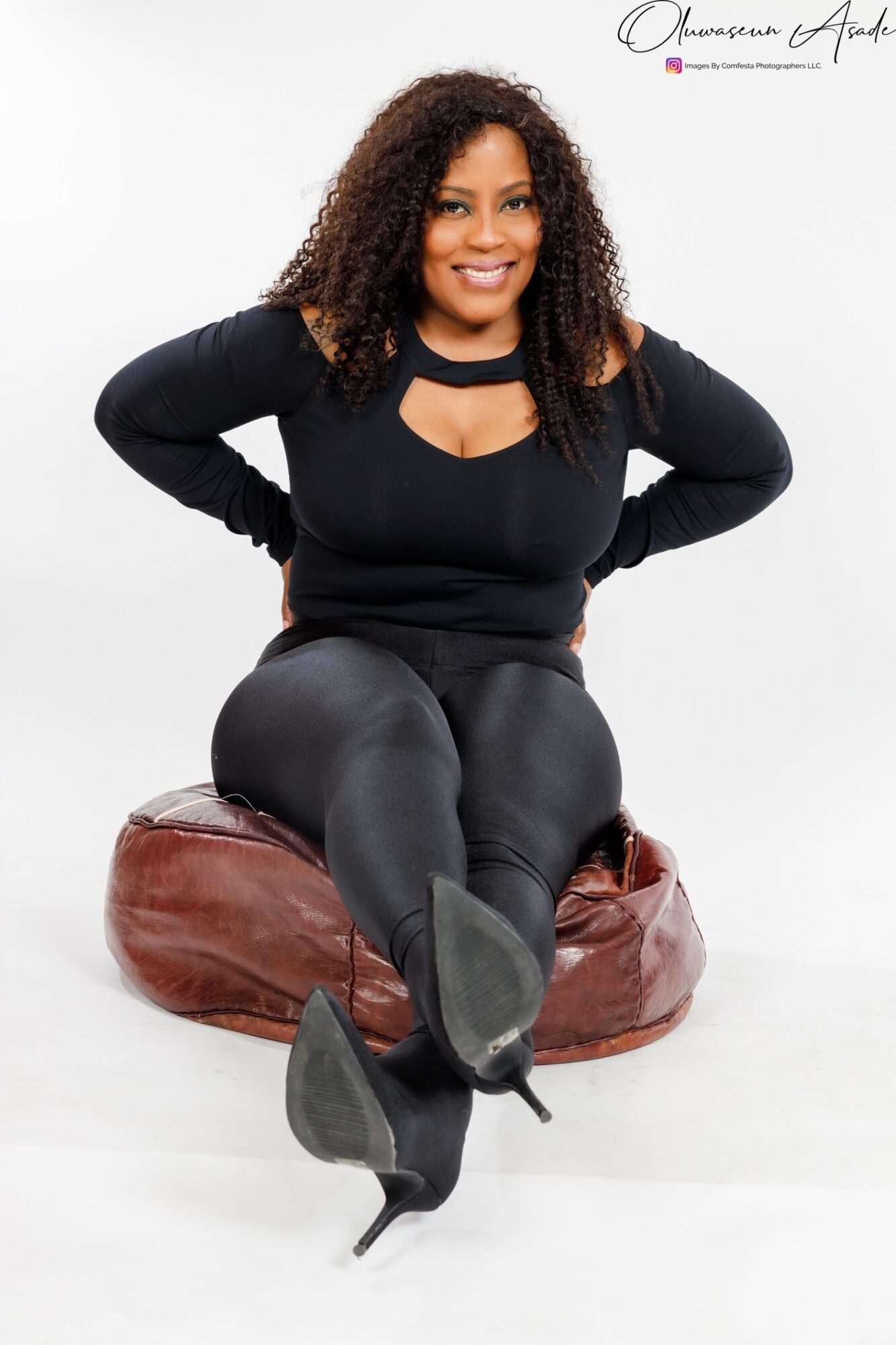 Woman sitting on a brown cushion, smiling, wearing a black top and pants, with curly hair, hands on hips.