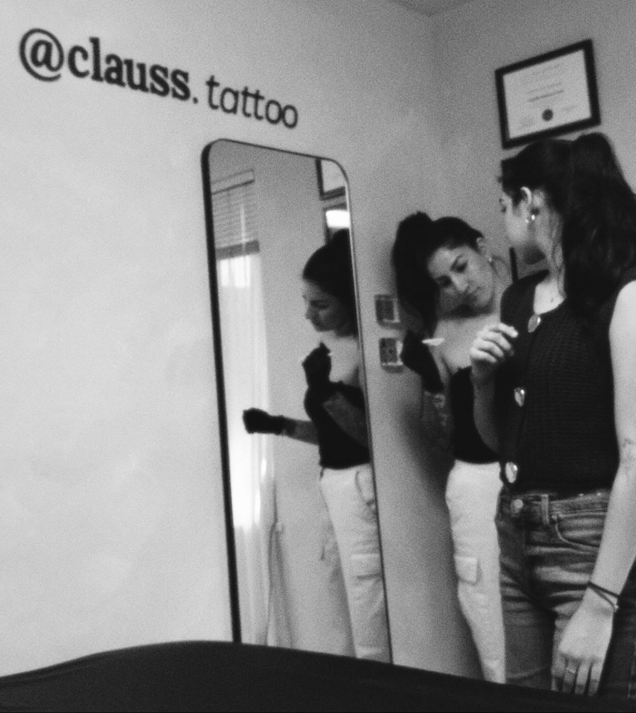 Two women stand near a mirror, one touching her face, in a tattoo studio with a framed certificate on the wall.