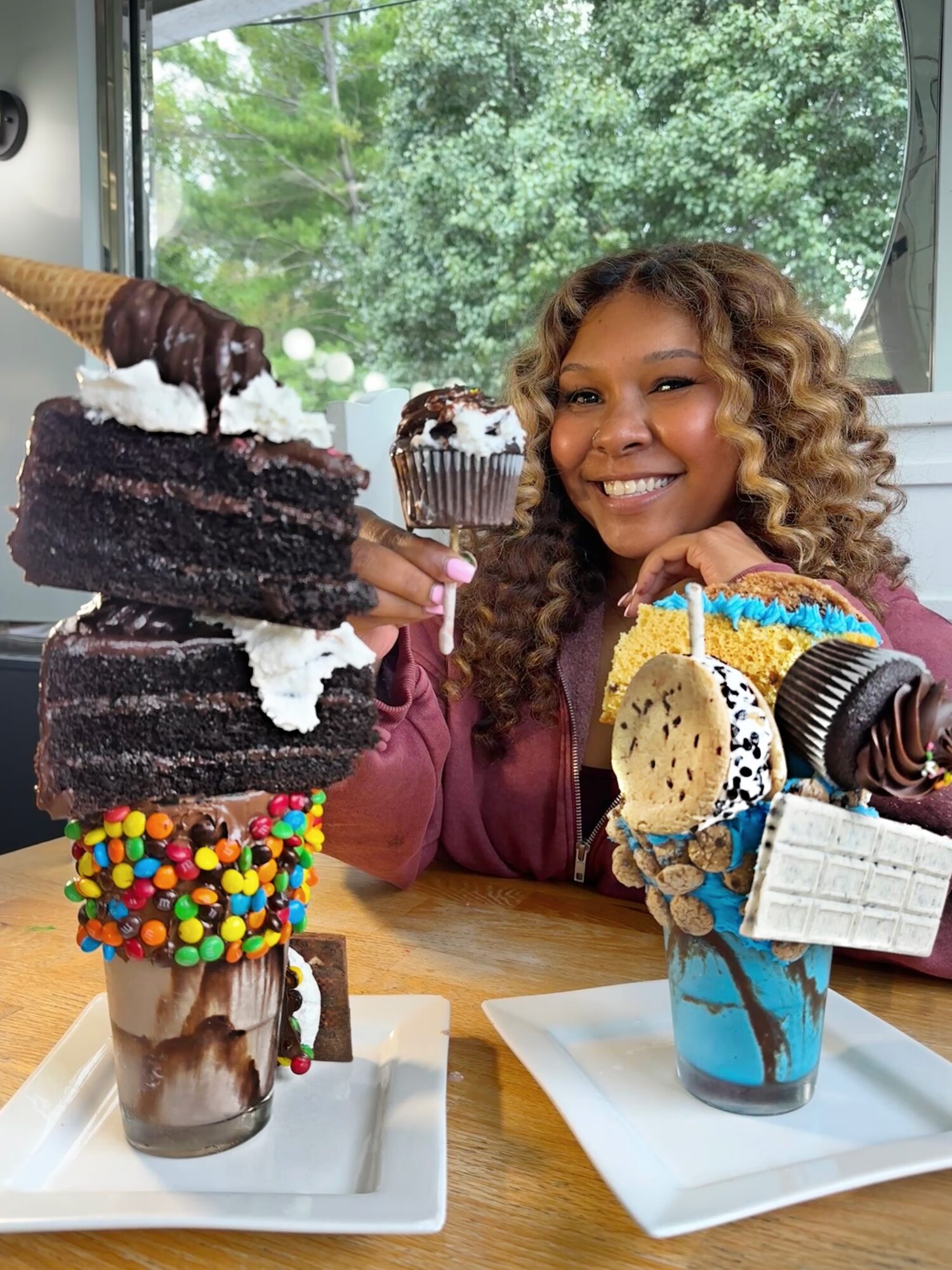 Smiling woman with curly hair sitting at a table with various desserts and ice cream, with a window and green trees outside.