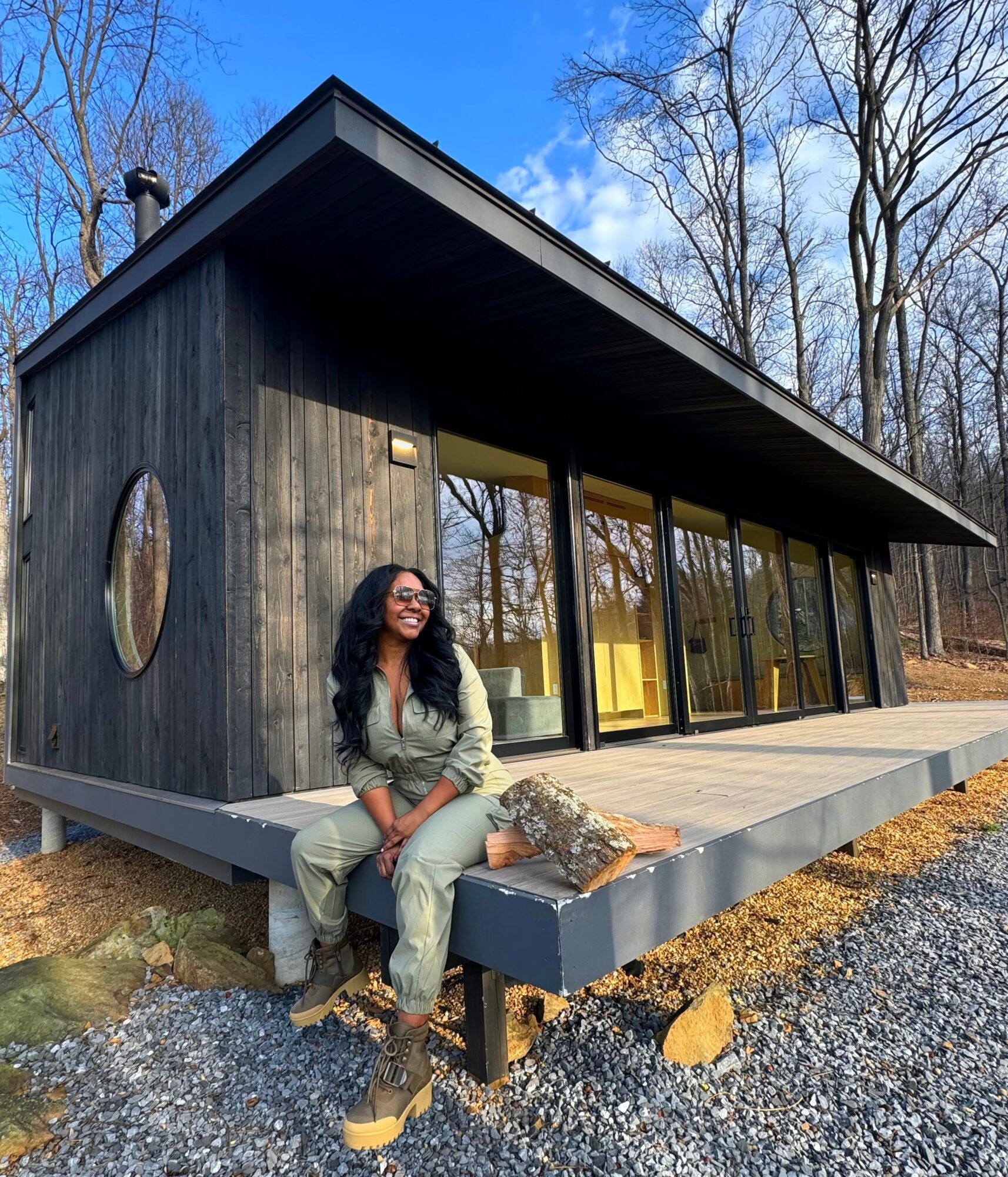 Woman sitting on a raised platform outside a modern house with large glass windows, surrounded by trees and gravel.