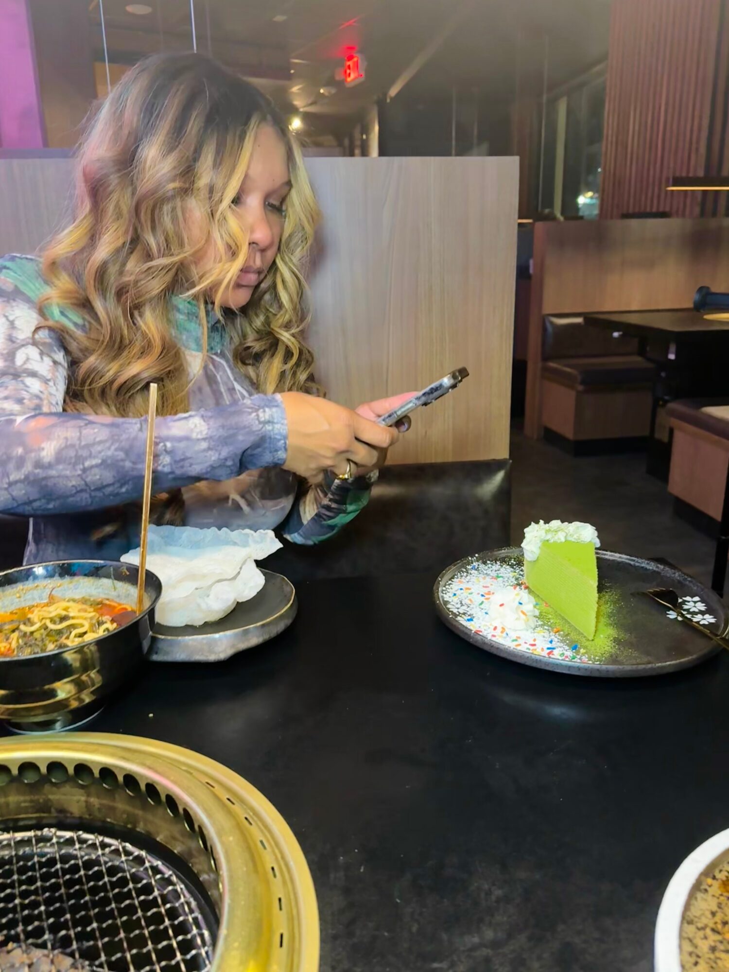 Woman with wavy blonde hair looking at her phone in a restaurant, with a slice of green cake on a plate nearby.