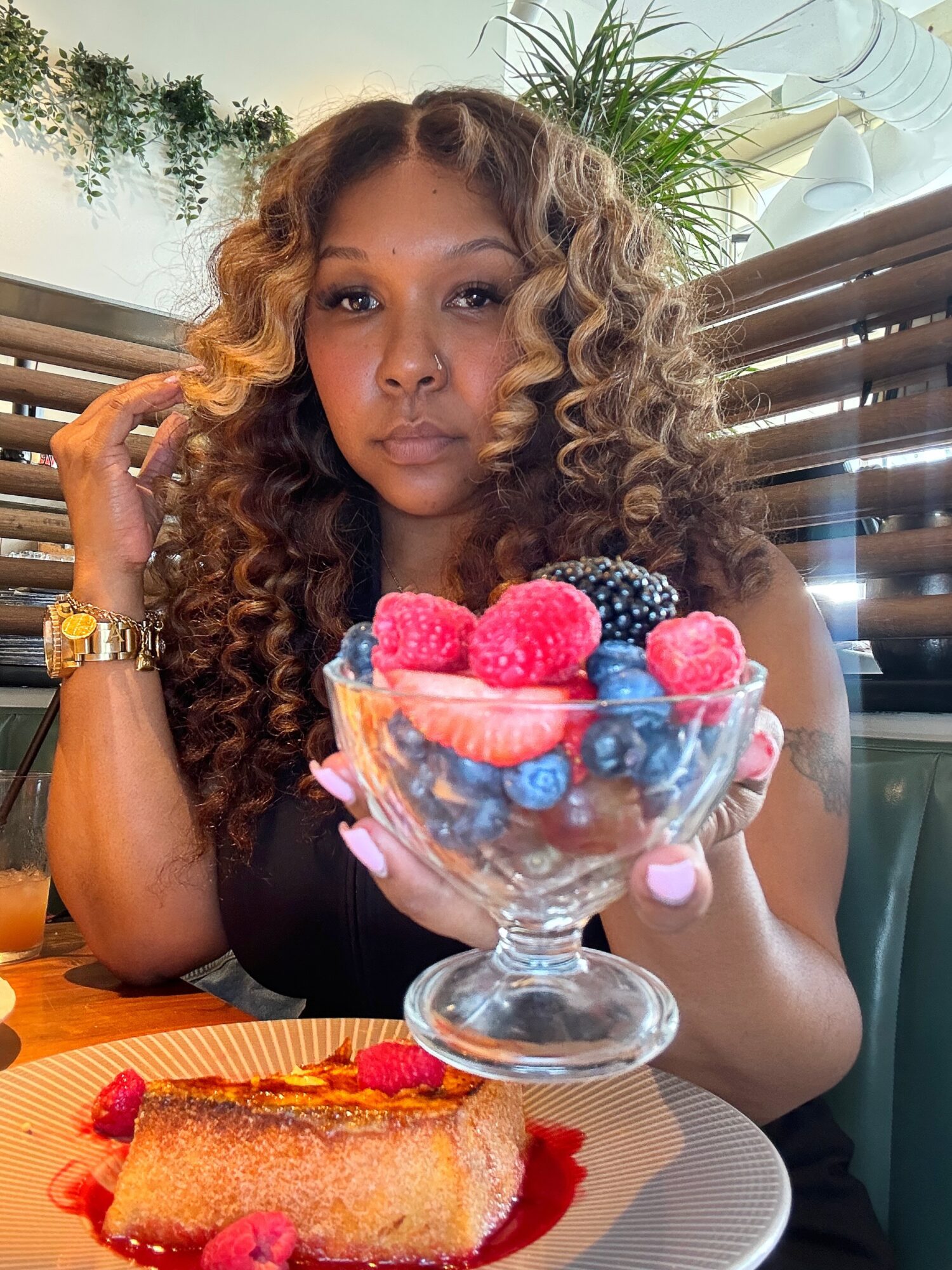 Woman with curly hair holding a glass bowl of berries and a slice of cake with raspberry sauce, sitting at a table.