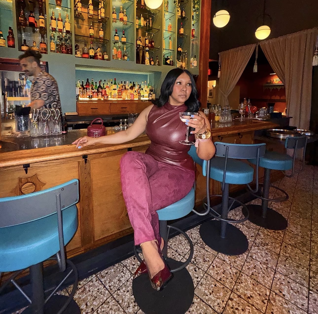 Woman sitting at bar holding a drink, bar shelves with bottles behind her, dim lighting, blue chairs, tiled floor.