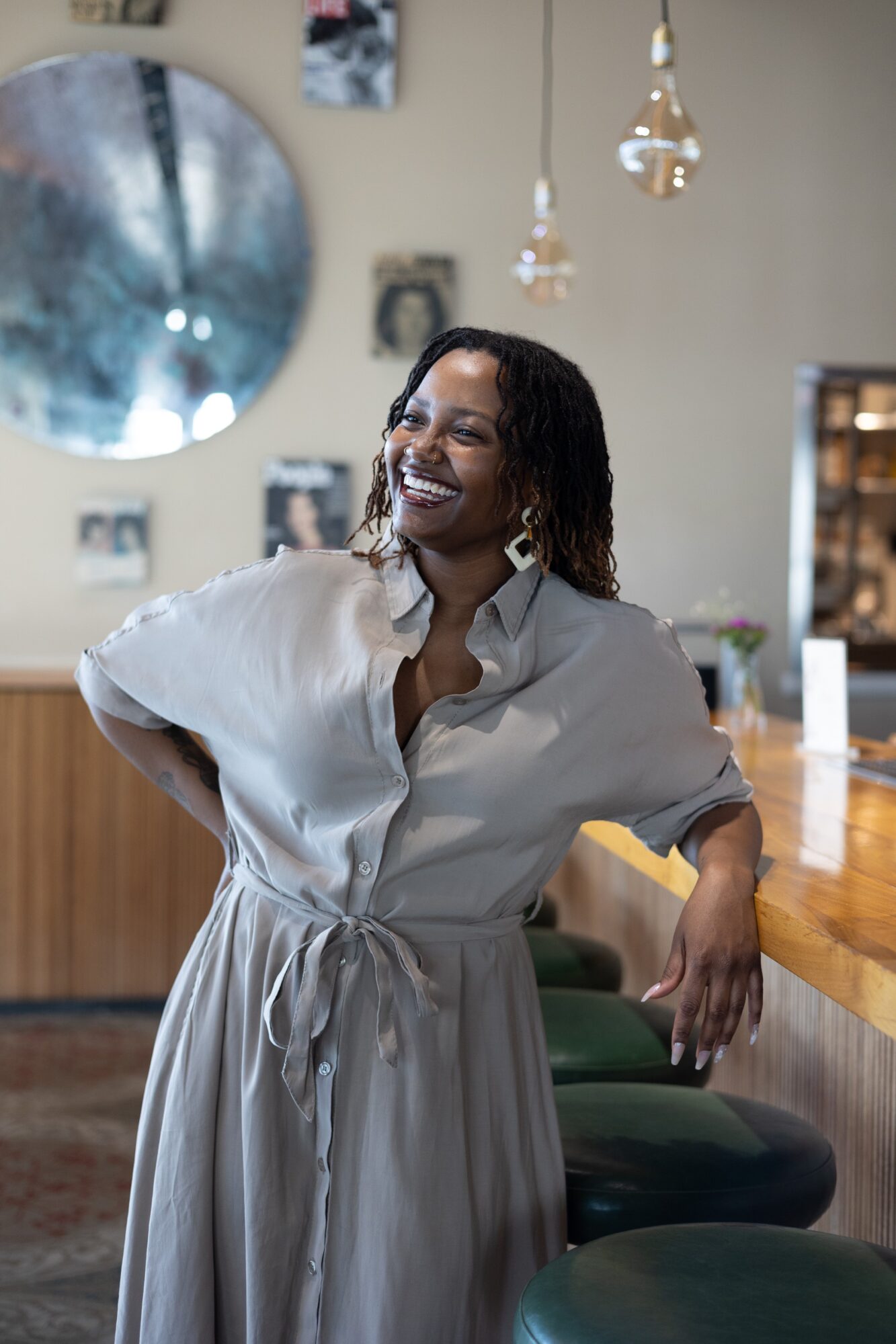 Woman with curly hair smiling, wearing a light-colored dress, standing in a modern cafe or restaurant interior.