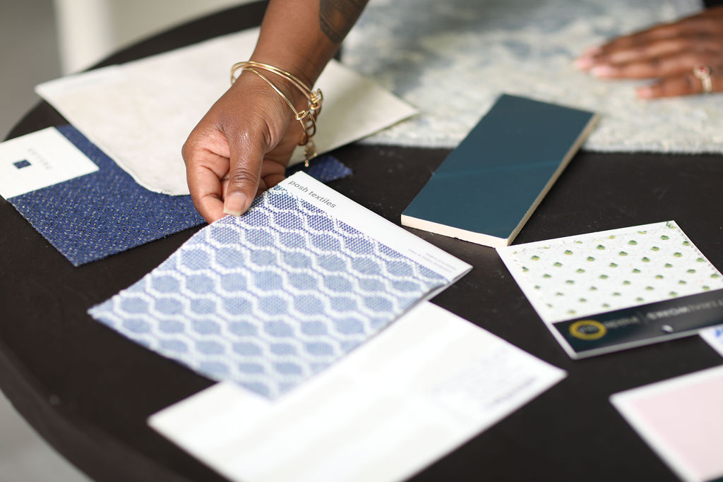 Person's hand holding a fabric sample, with various fabric swatches and papers on a table.