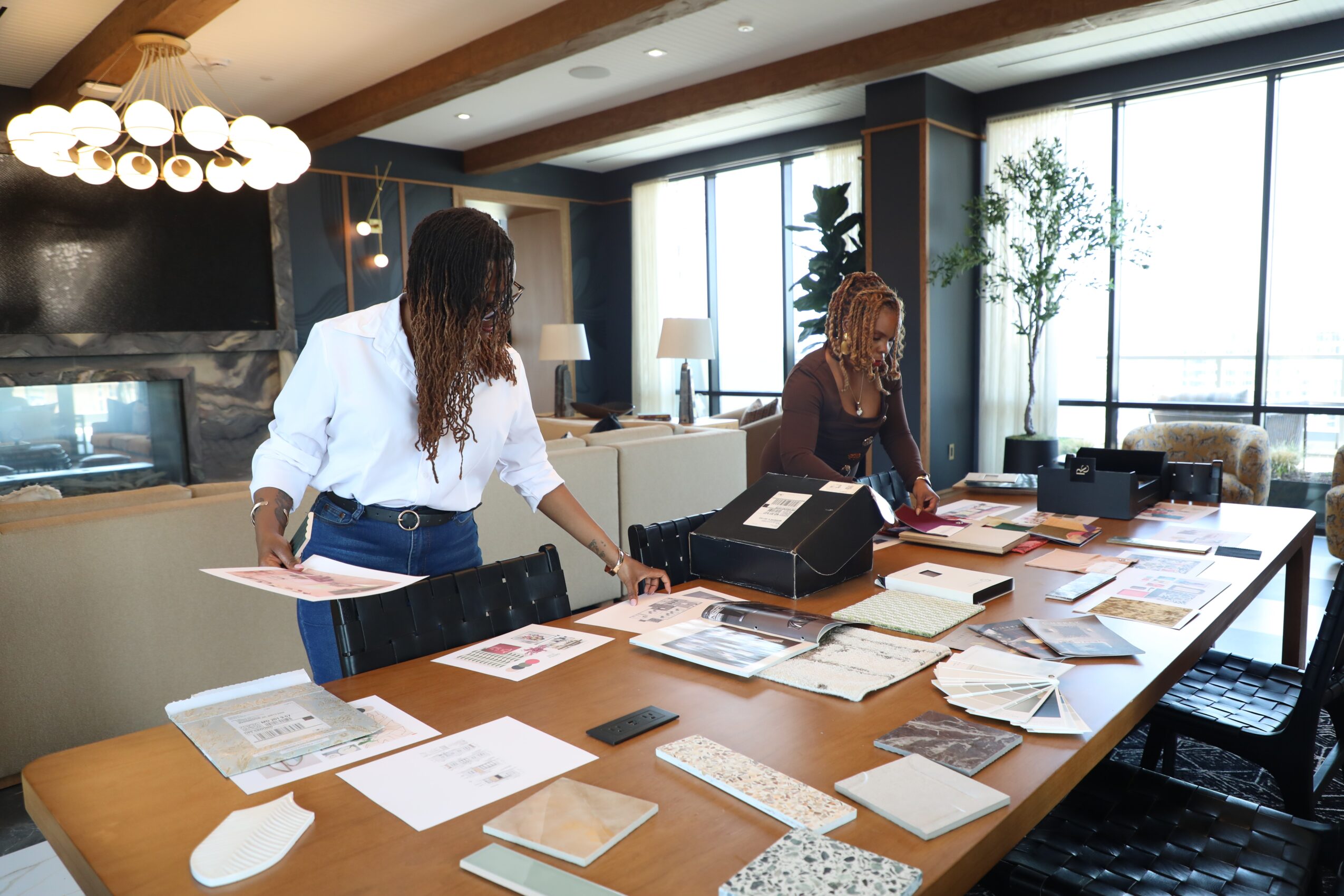 Two women stand at a large table with papers and folders in a modern, well-lit room with large windows and plants.
