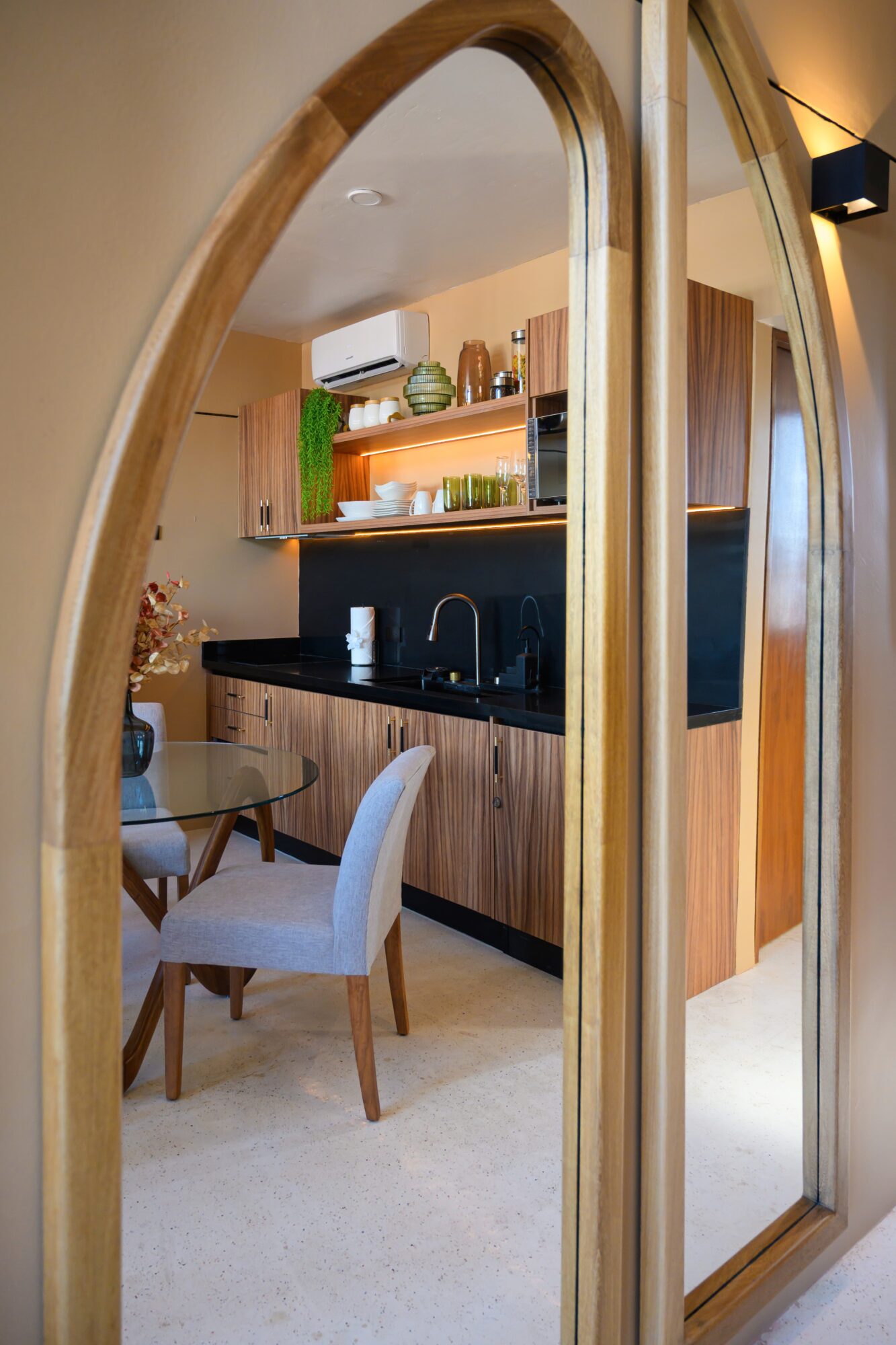 View through an arched mirror showing a kitchen with wooden cabinets, black countertop, and a dining area with chairs.