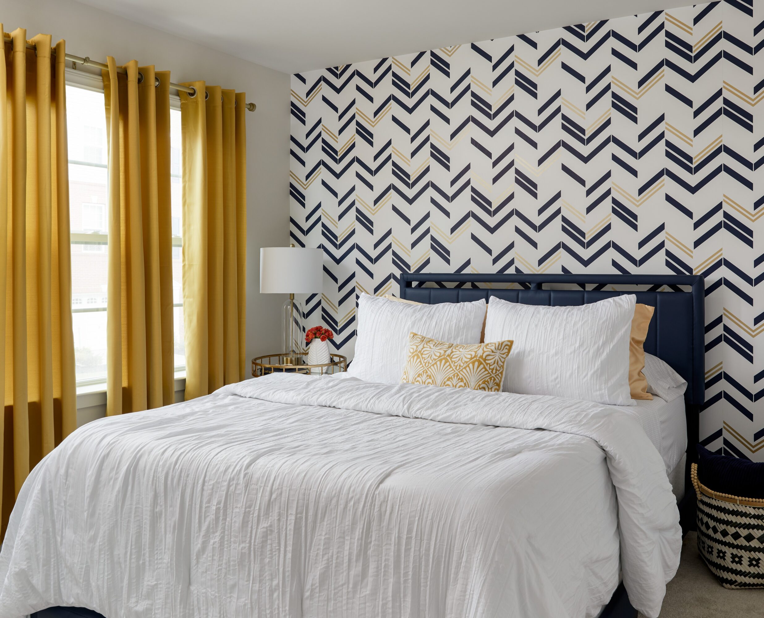 Bedroom with a bed, white bedding, decorative pillow, yellow curtains, patterned accent wall, and a nightstand with a lamp.
