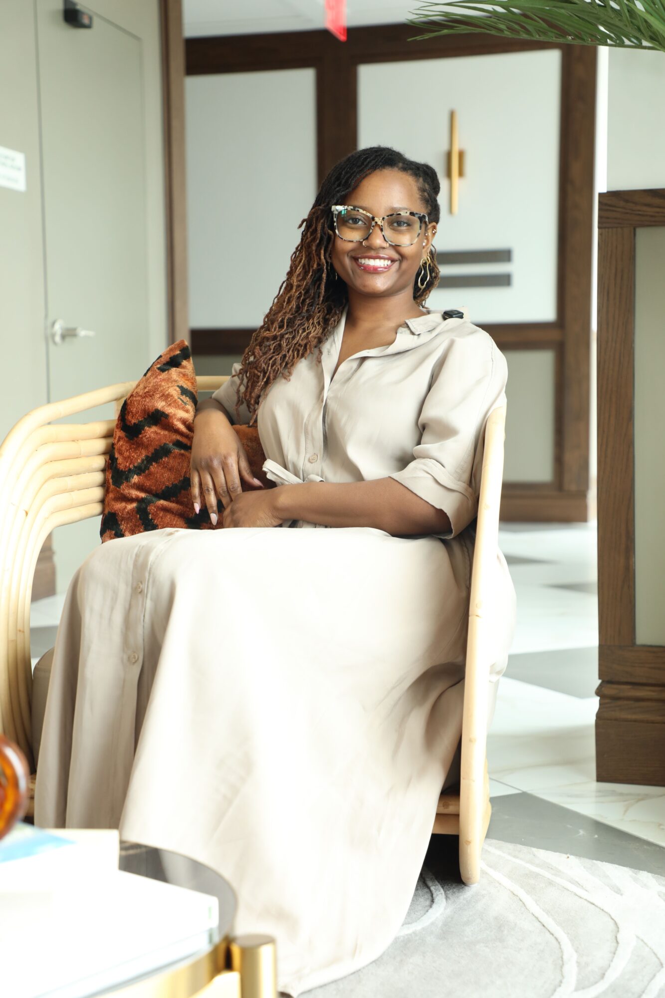 Woman with glasses and long hair sitting on a chair, smiling, in a room with a door and a plant.