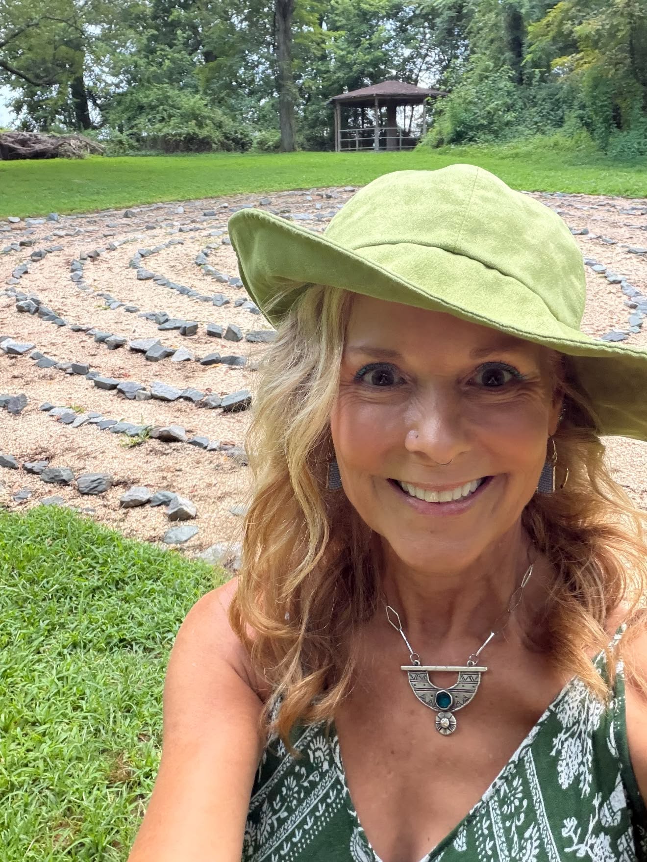 Woman smiling outdoors with a large green hat, wearing a patterned dress and necklace, in a garden with a labyrinth and gazebo.