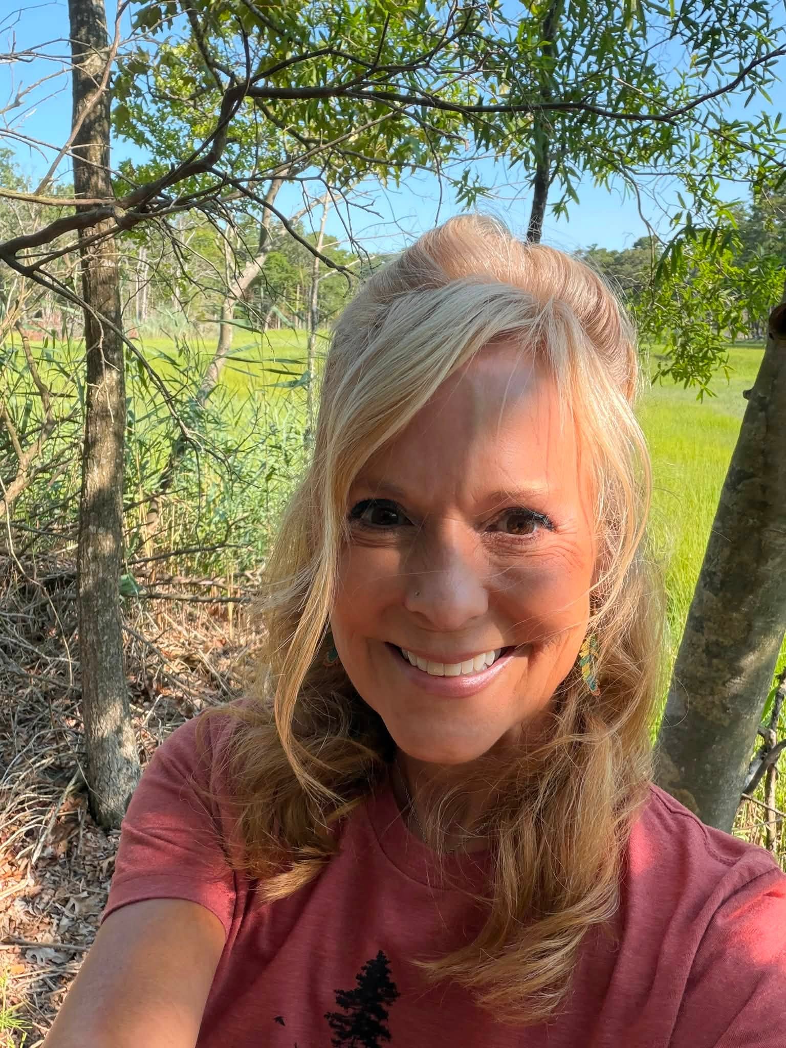 Smiling woman with blonde hair outdoors, trees and greenery in background.