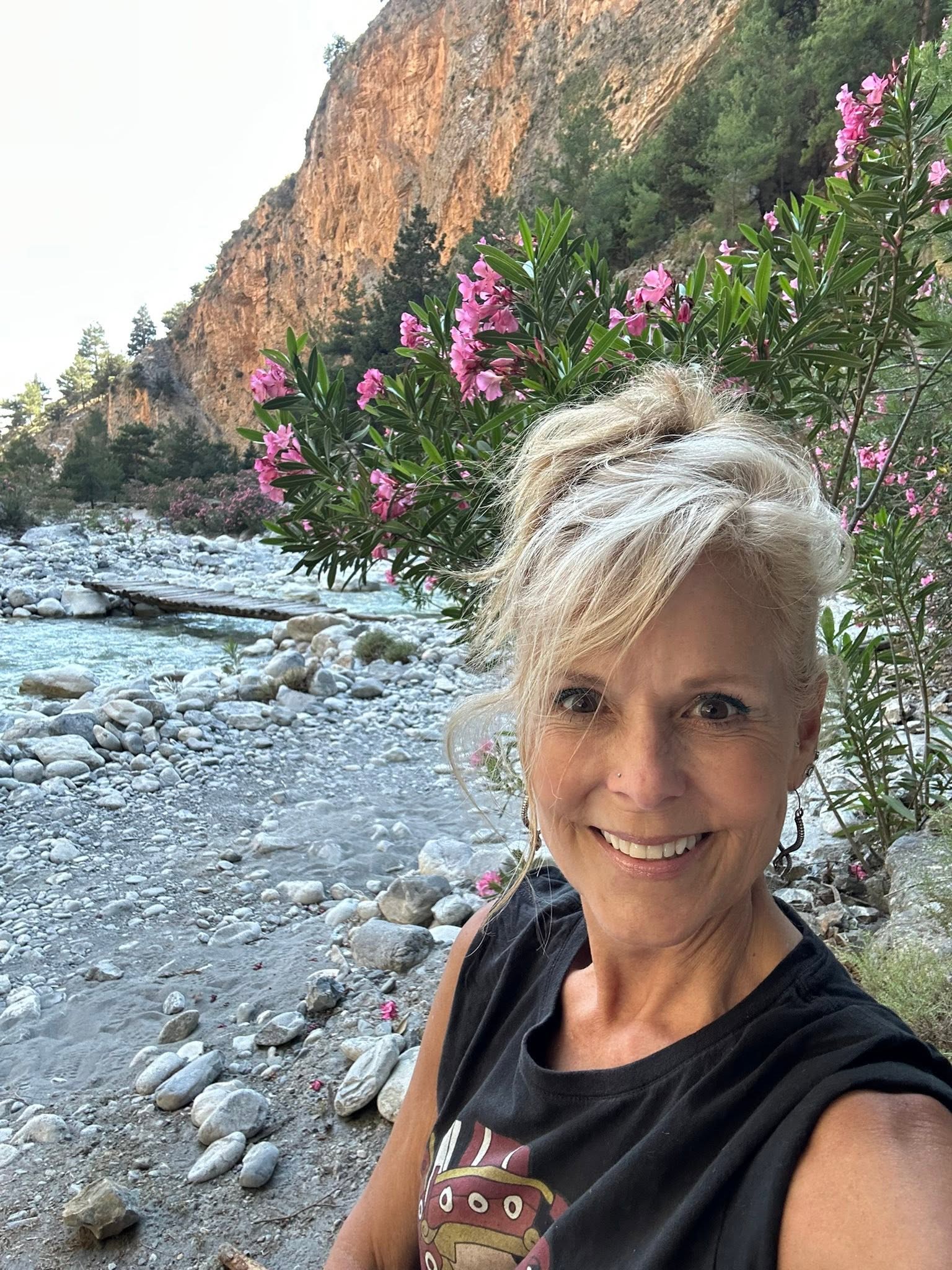 Smiling woman with short blonde hair outdoors near pink flowering bushes and rocky river, mountain in background.