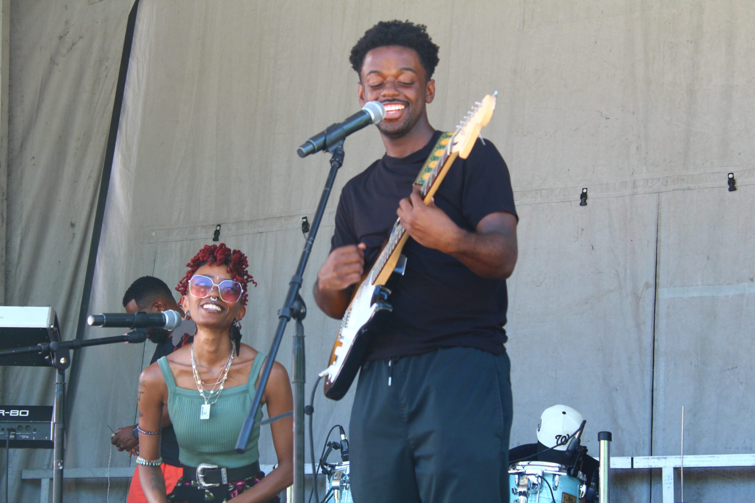 Two musicians perform on stage, one singing into a microphone and playing guitar, the other smiling with sunglasses and jewelry.