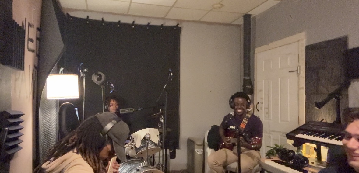 People in a music studio with microphones, instruments, and recording equipment, some sitting and some standing, with a black curtain backdrop.