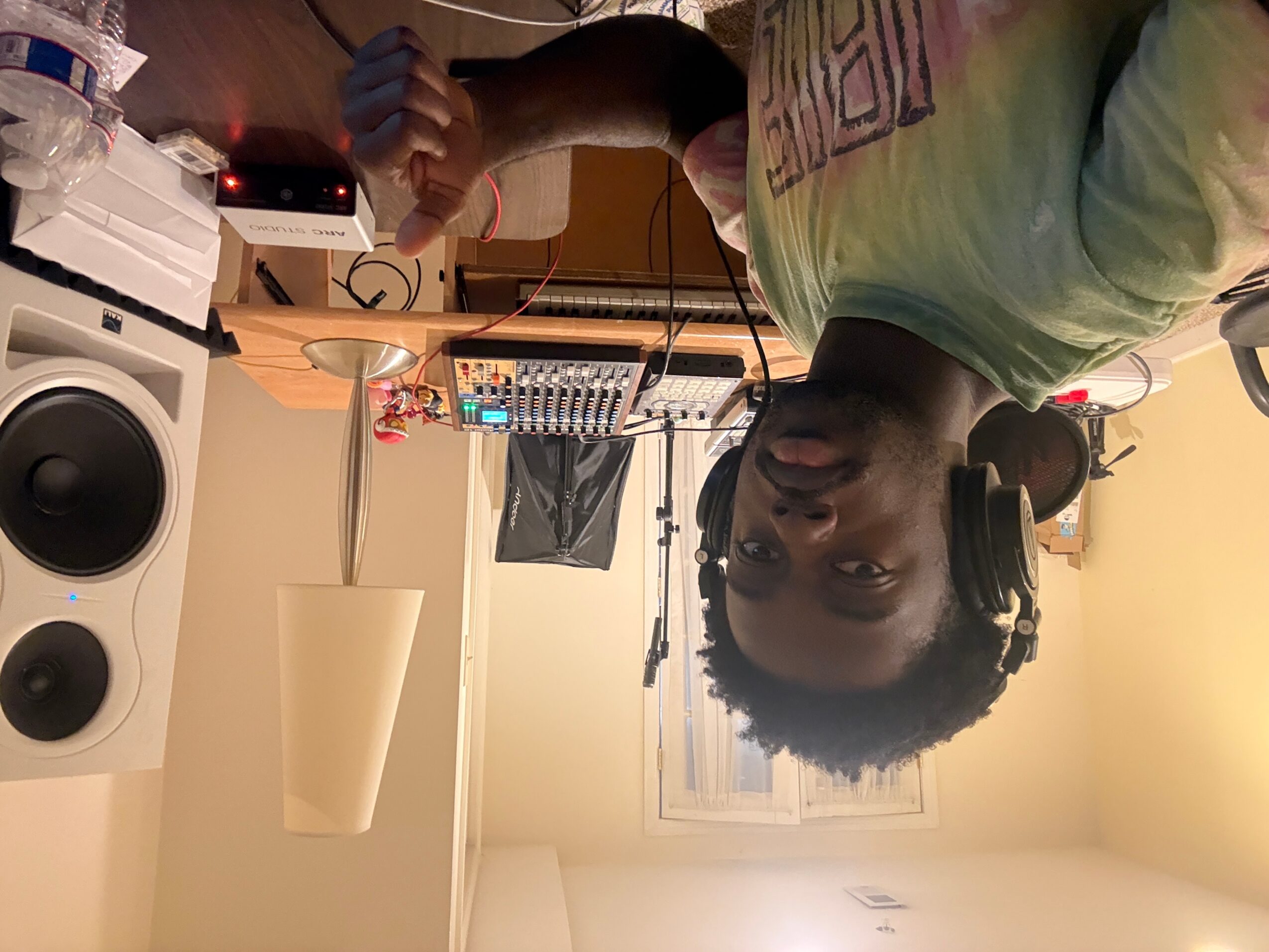 Person wearing headphones in a room with audio equipment, a speaker, and a window, facing the camera.