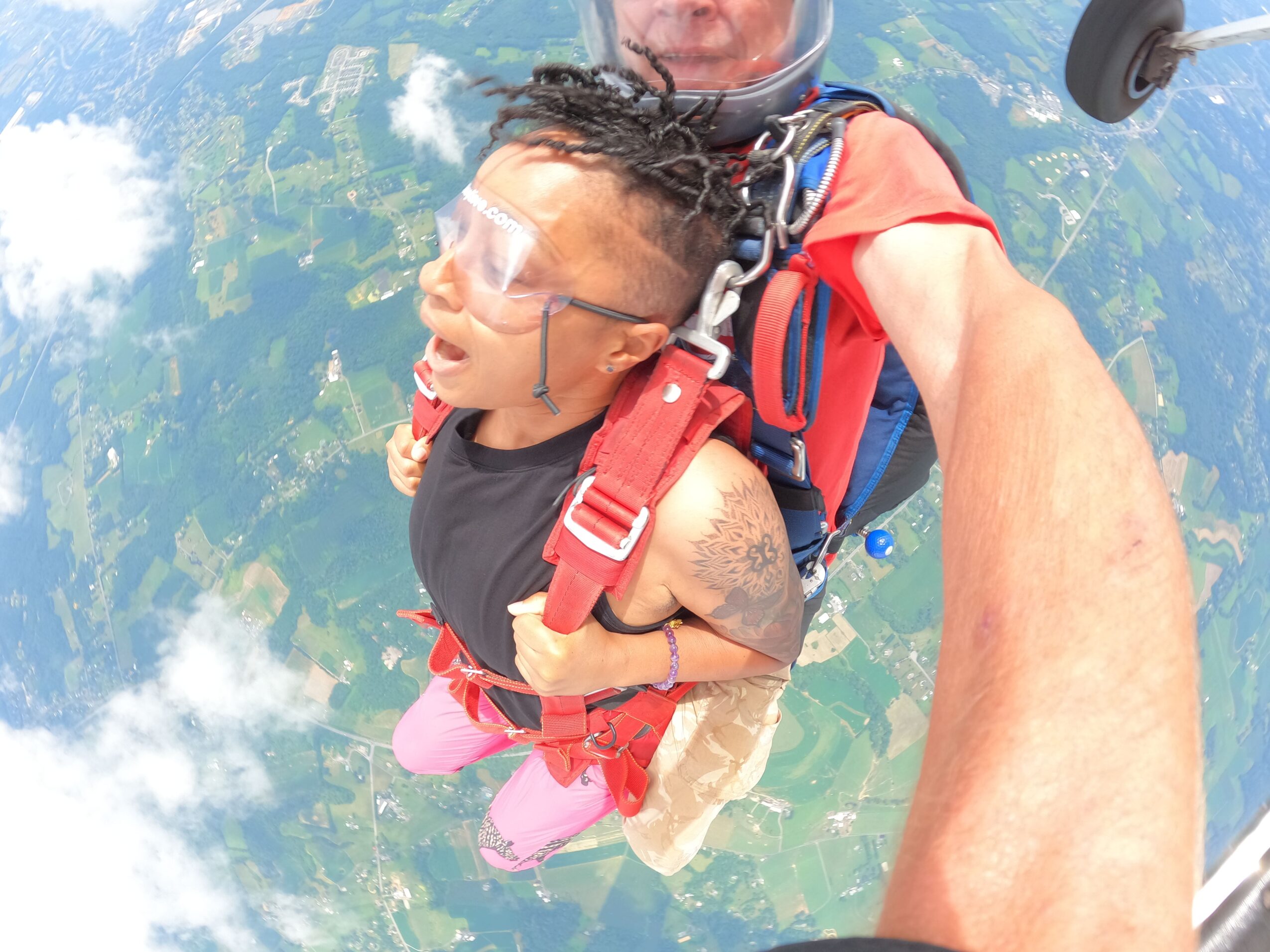 Person skydiving with instructor, open sky and landscape below, person wearing goggles and harness, instructor holding person securely.