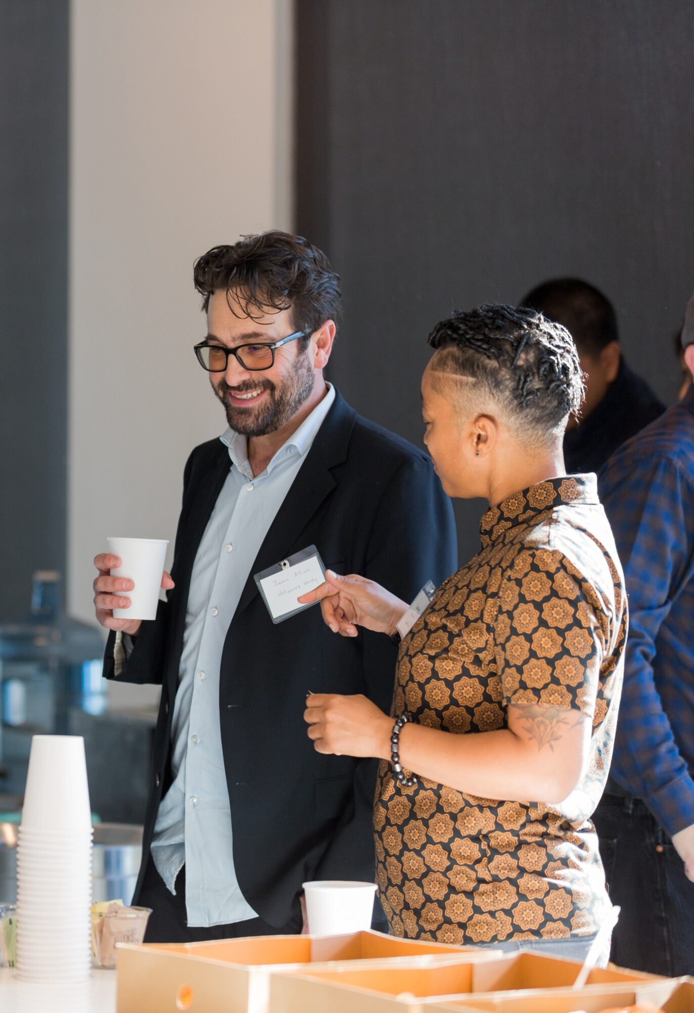 Two people smiling and talking, one holding a cup, in a professional setting with others in the background.
