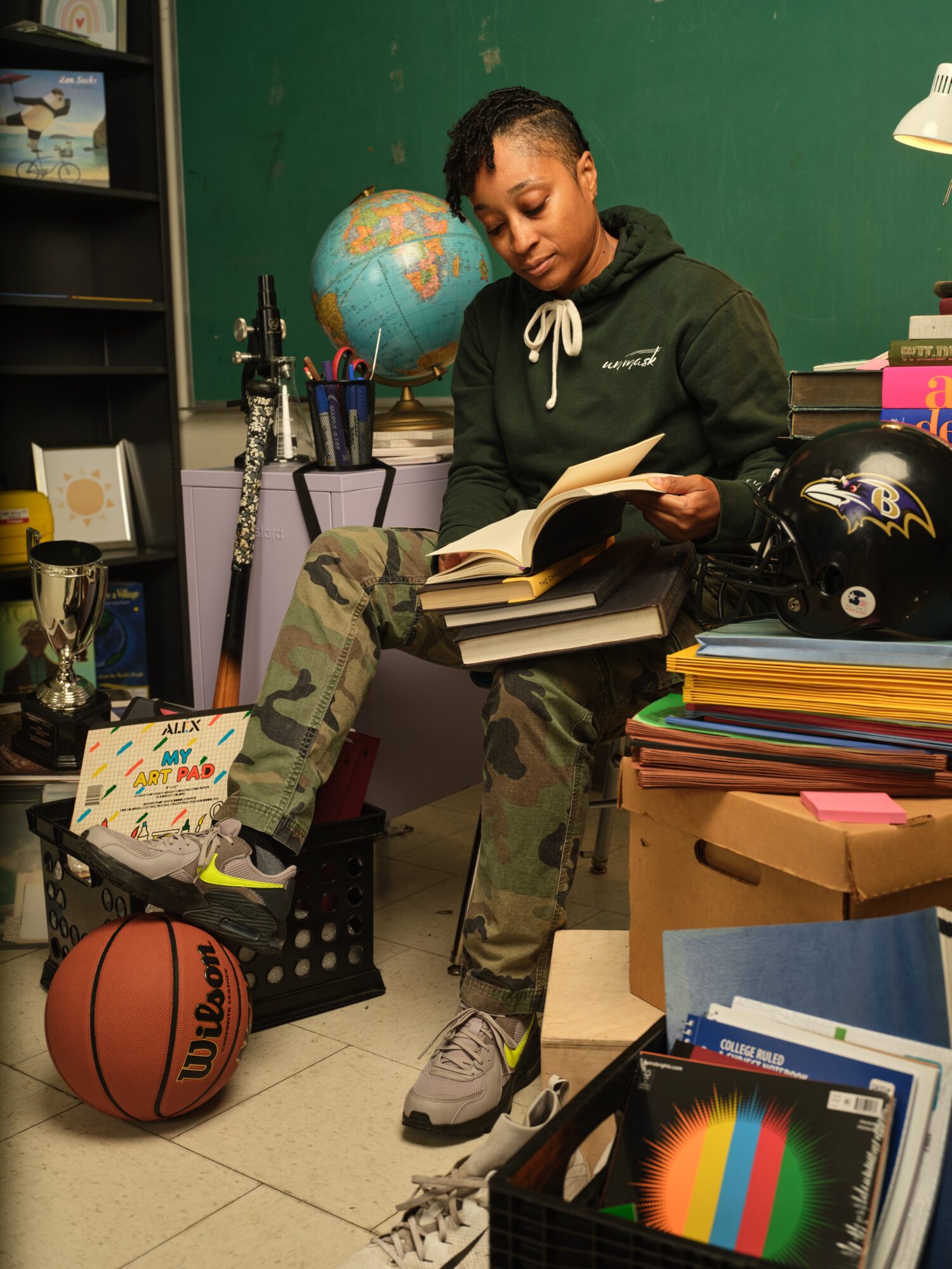 Person sitting on a chair reading a book in a classroom with a globe, sports equipment, and books around.
