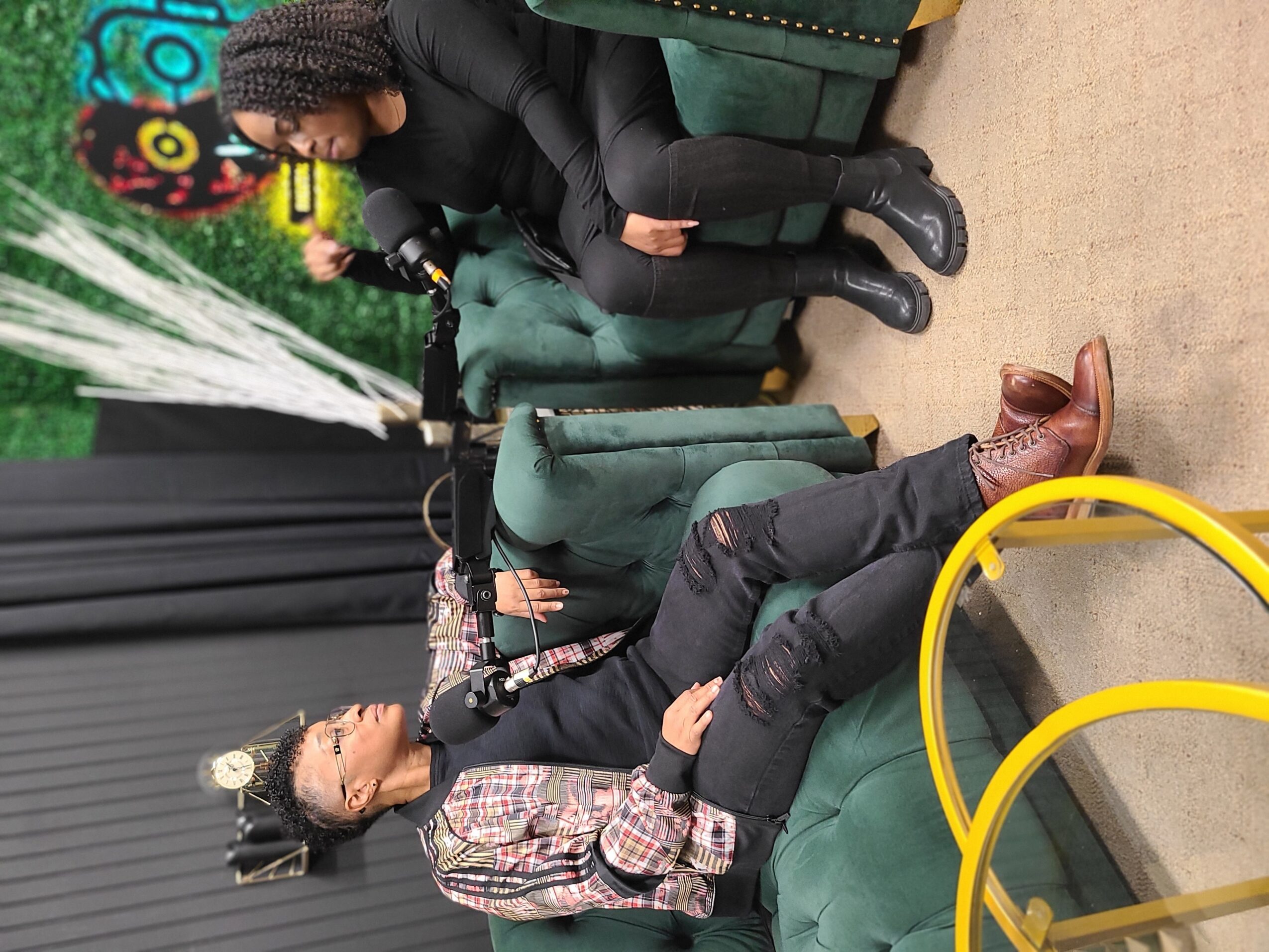 Two women sitting on green chairs in a room with black curtains and colorful wall art, one speaking into a microphone.