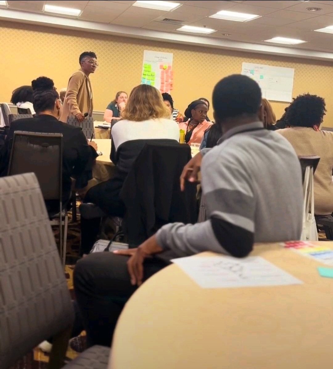 Group of people in a conference room, some seated and one standing, with presentation boards on the wall.