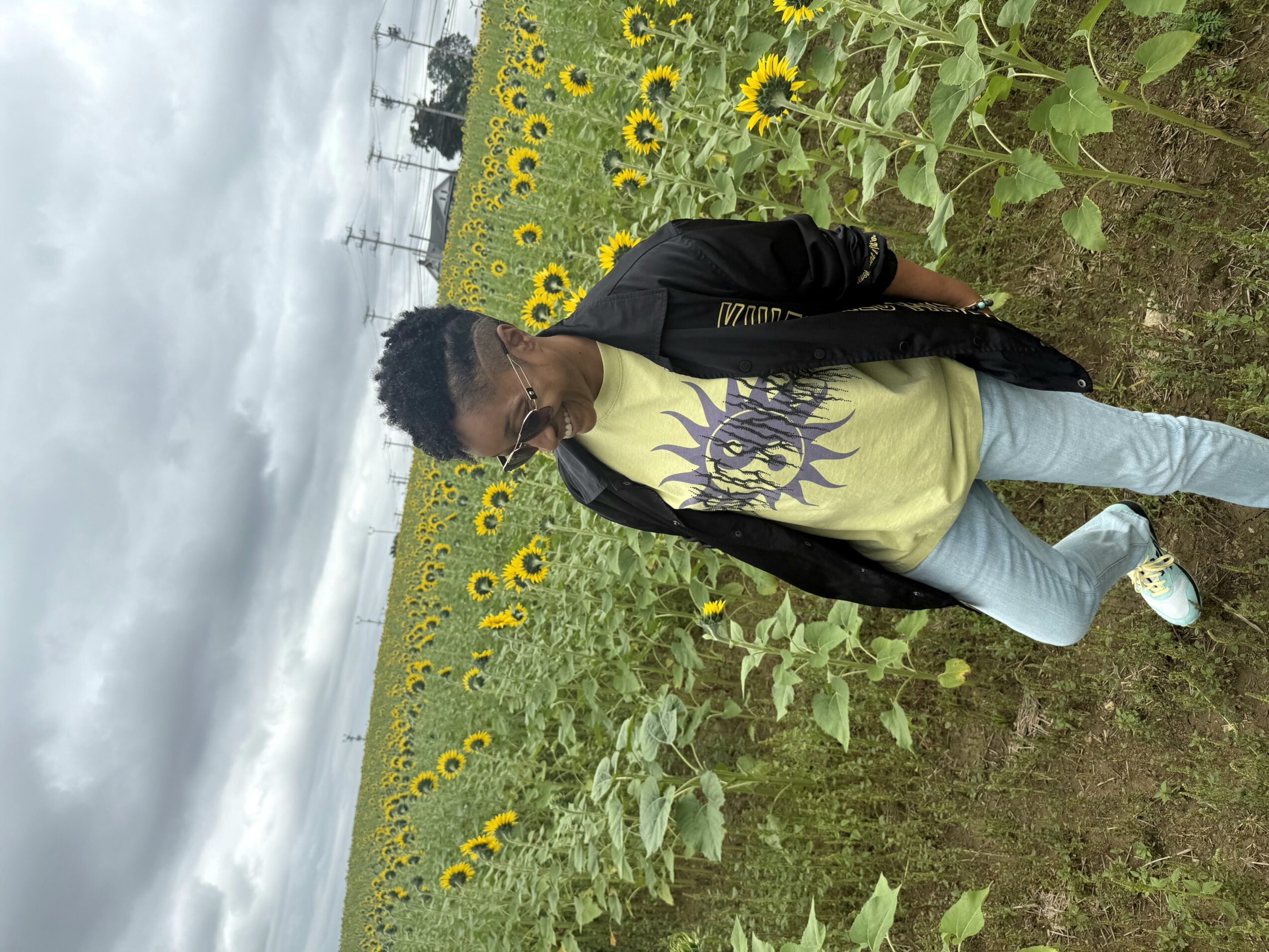 Person standing in a sunflower field with cloudy sky, wearing a black jacket, yellow shirt, and light blue jeans.