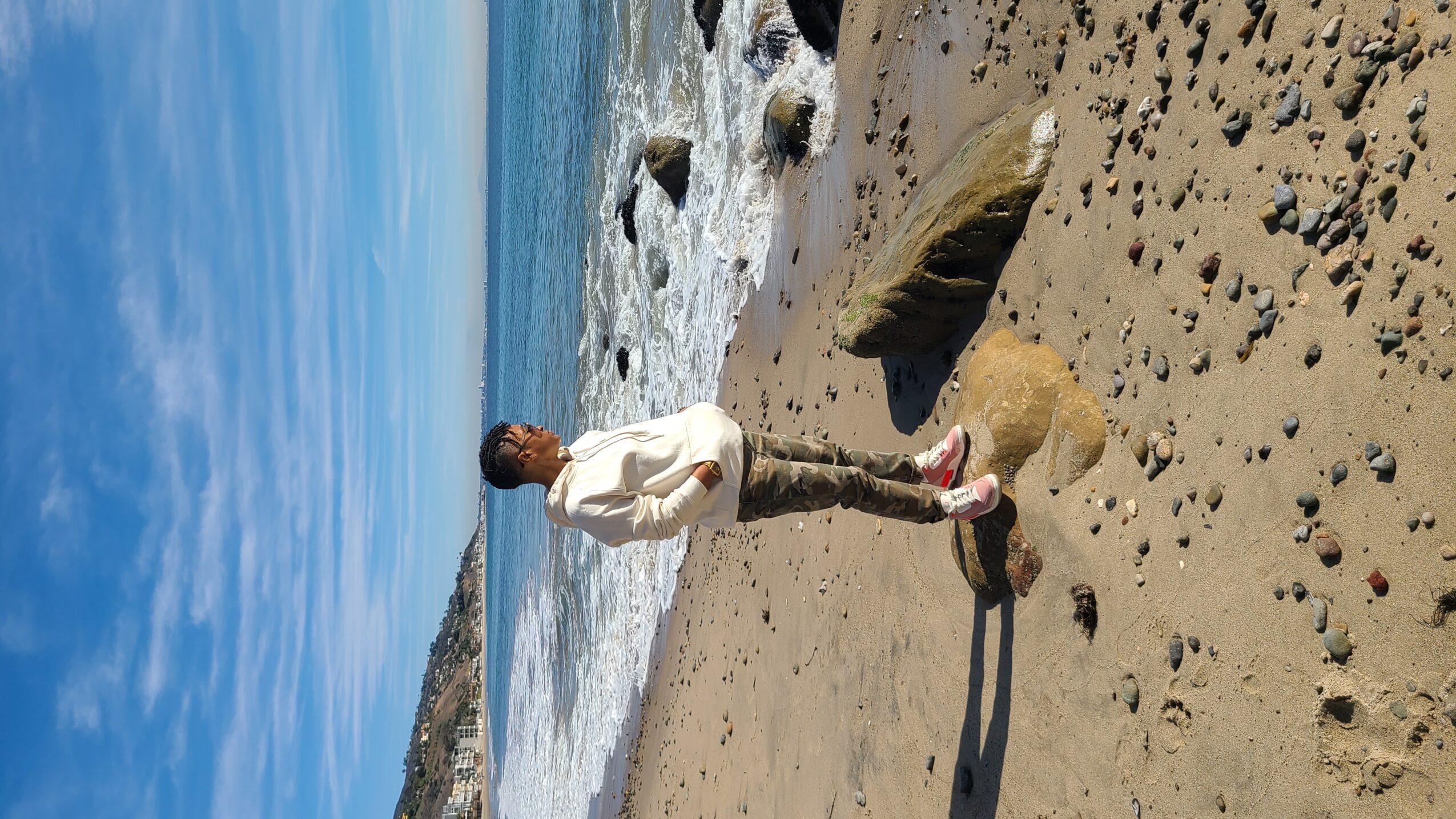 Person standing on sandy beach near rocks, looking at the ocean with blue sky and clouds, holding a dog leash.