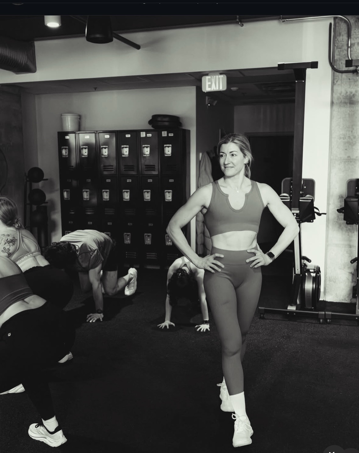 Woman in workout clothes standing with hands on hips in gym, with people exercising in background, lockers, and equipment visible.