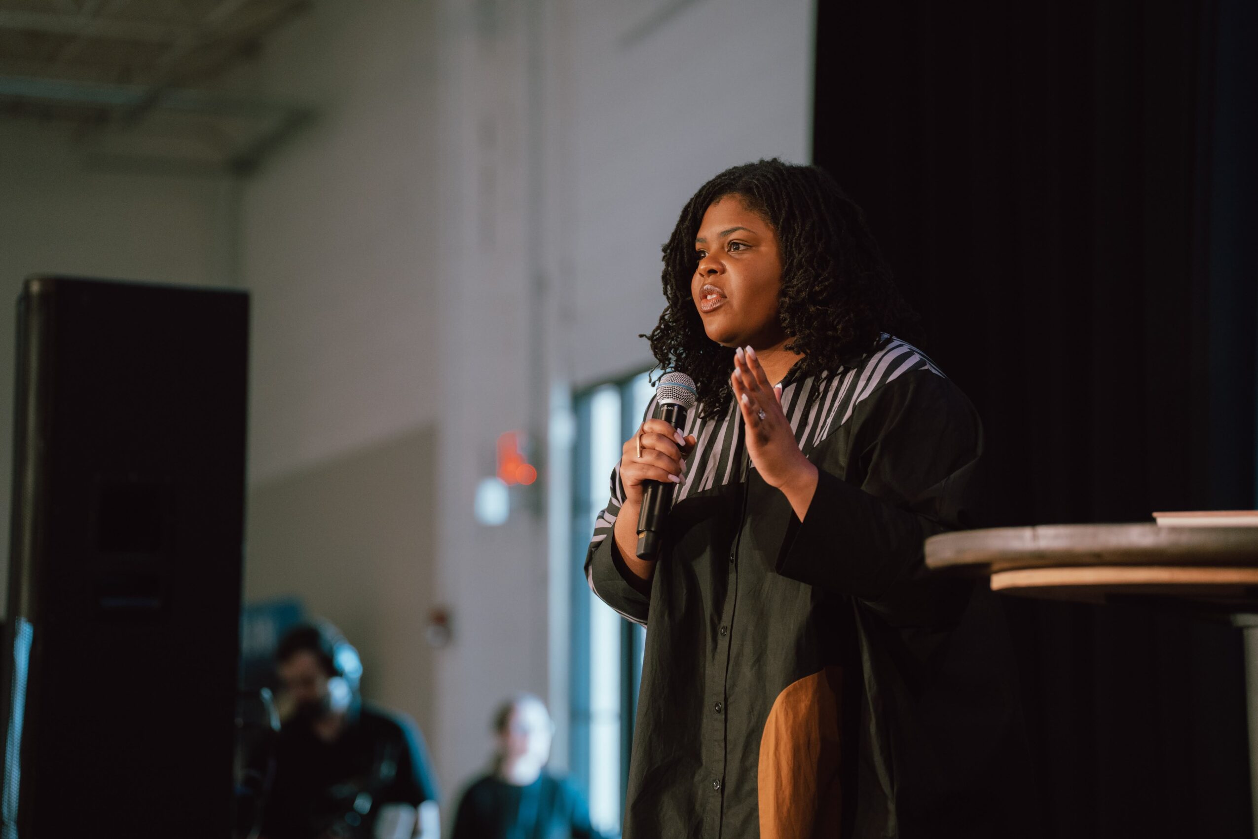 Woman speaking into microphone on stage, gesturing with hands, in a large indoor space with audience and equipment.